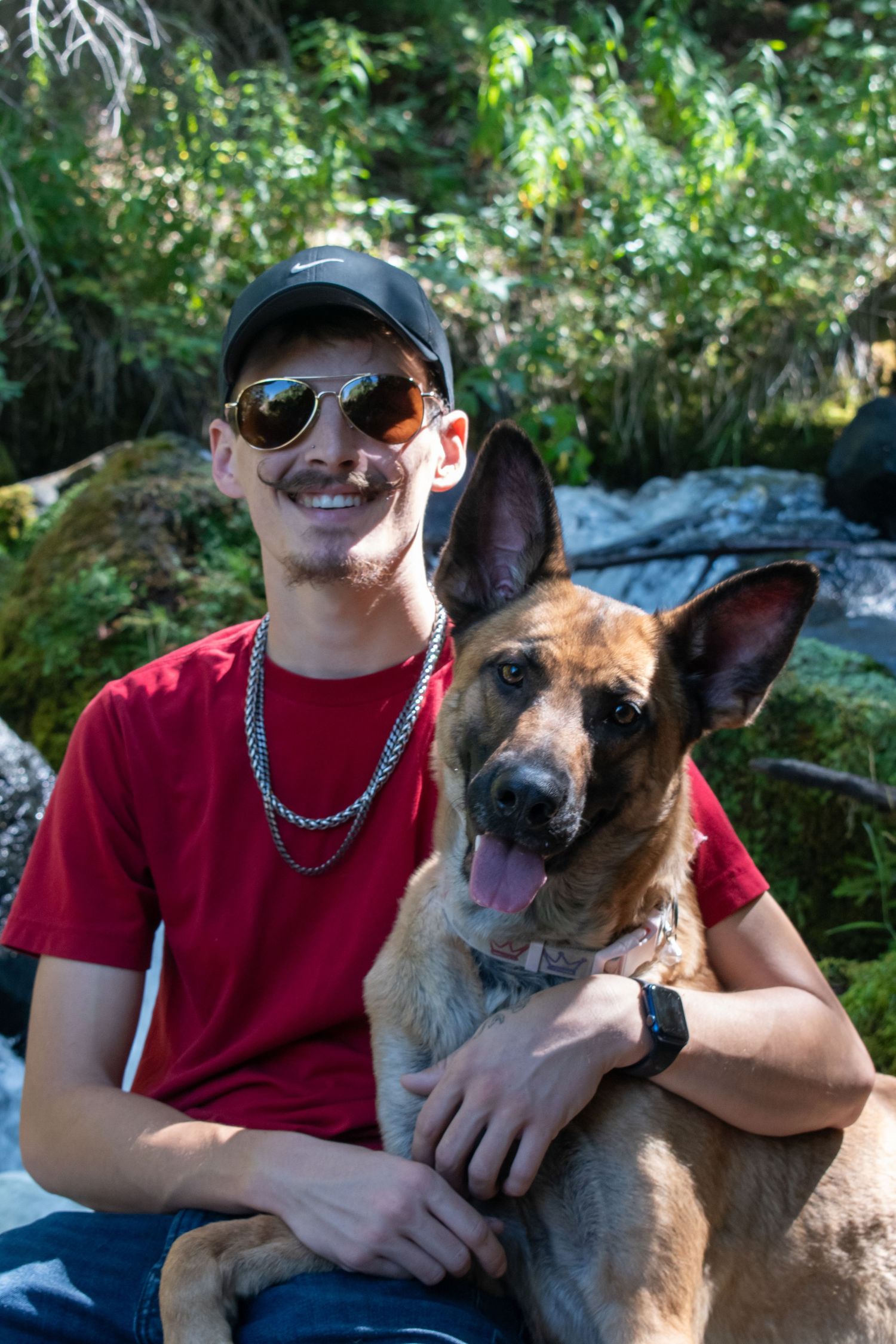 Young man in red shirt and sunglasses smiling with German Shepherd dog in forest setting.