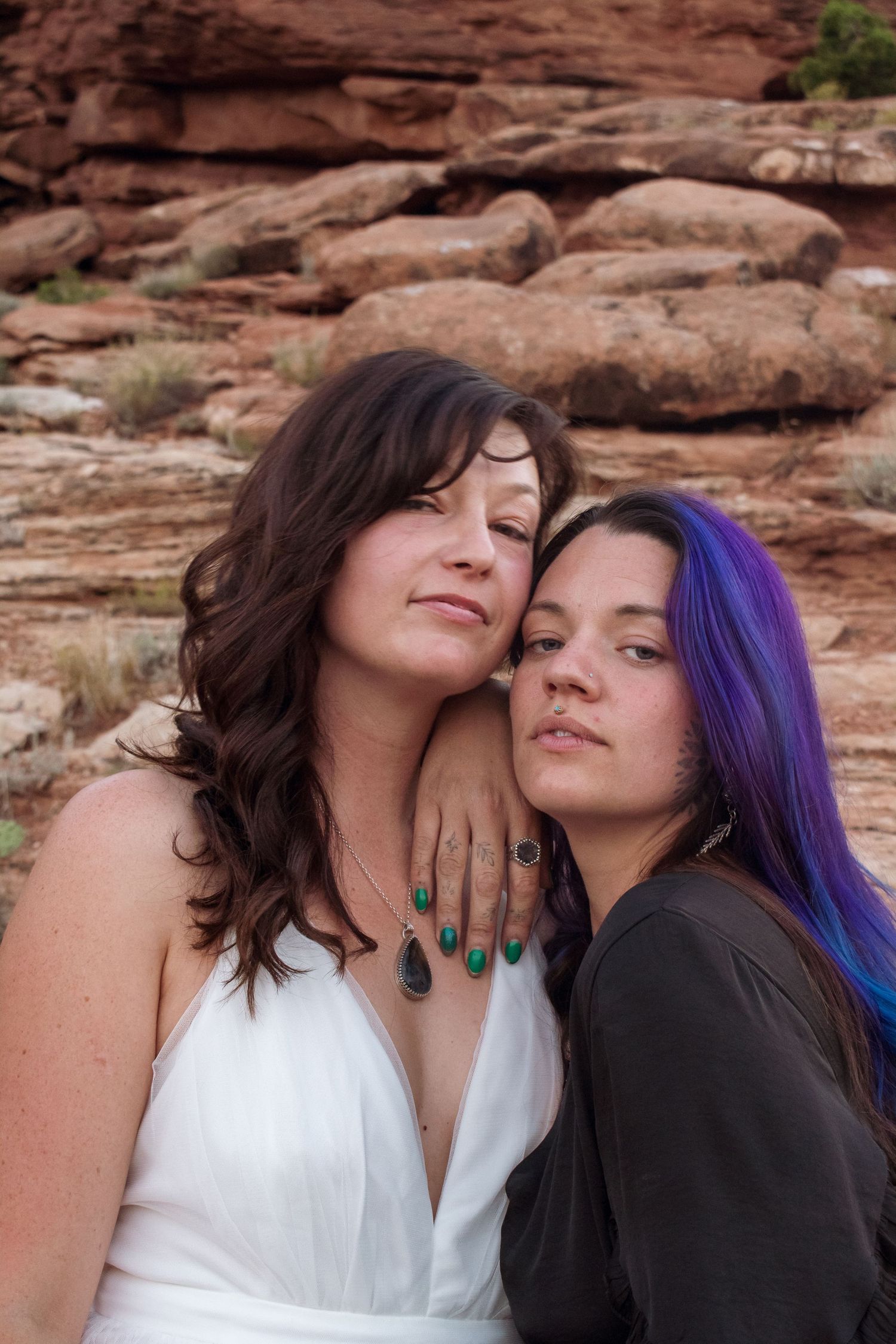 Two women pose together against red rock formations, one in white dress with green nails, other with purple hair.
