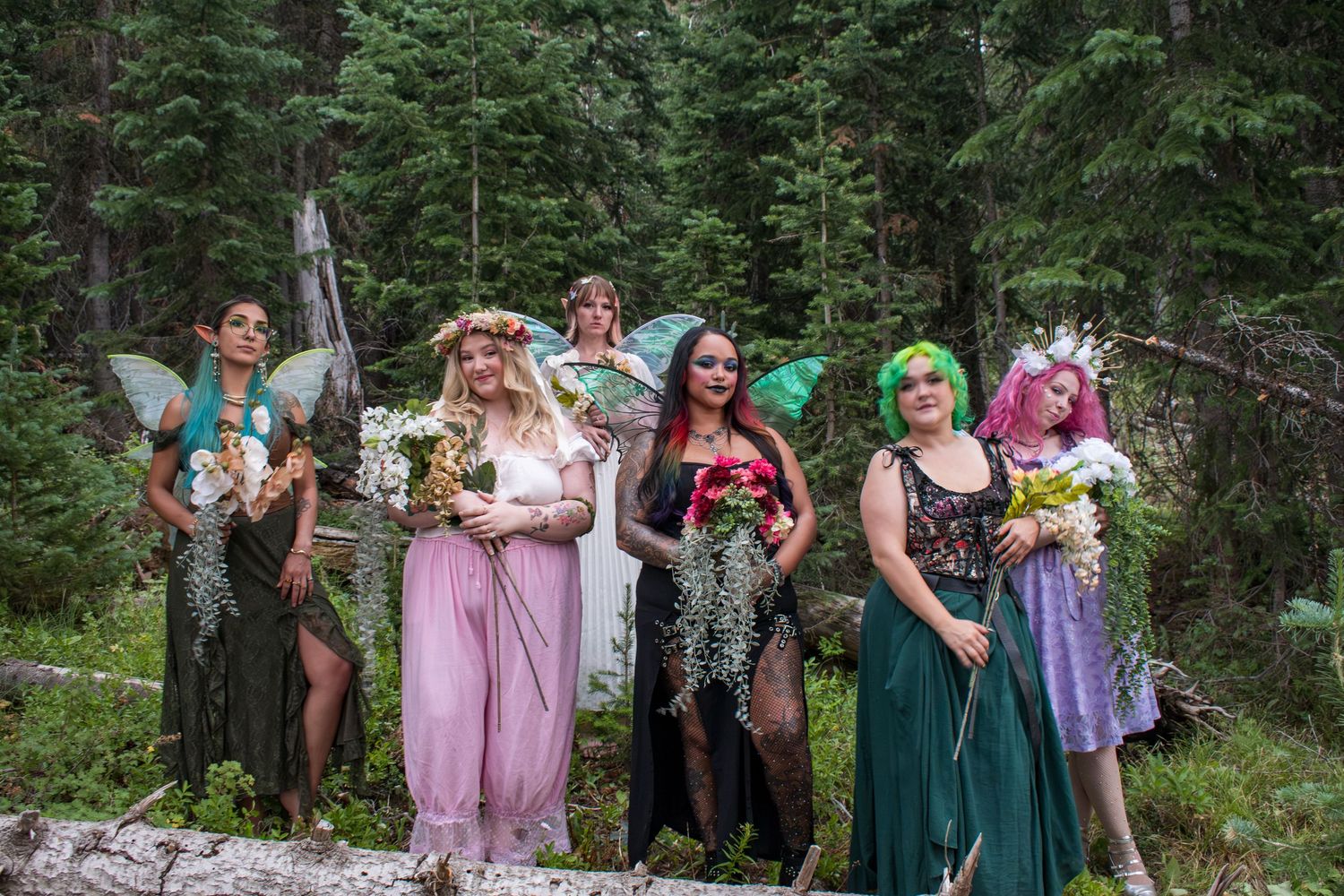 Group of women in fairy costumes with wings and flower crowns posing in a lush green forest setting.