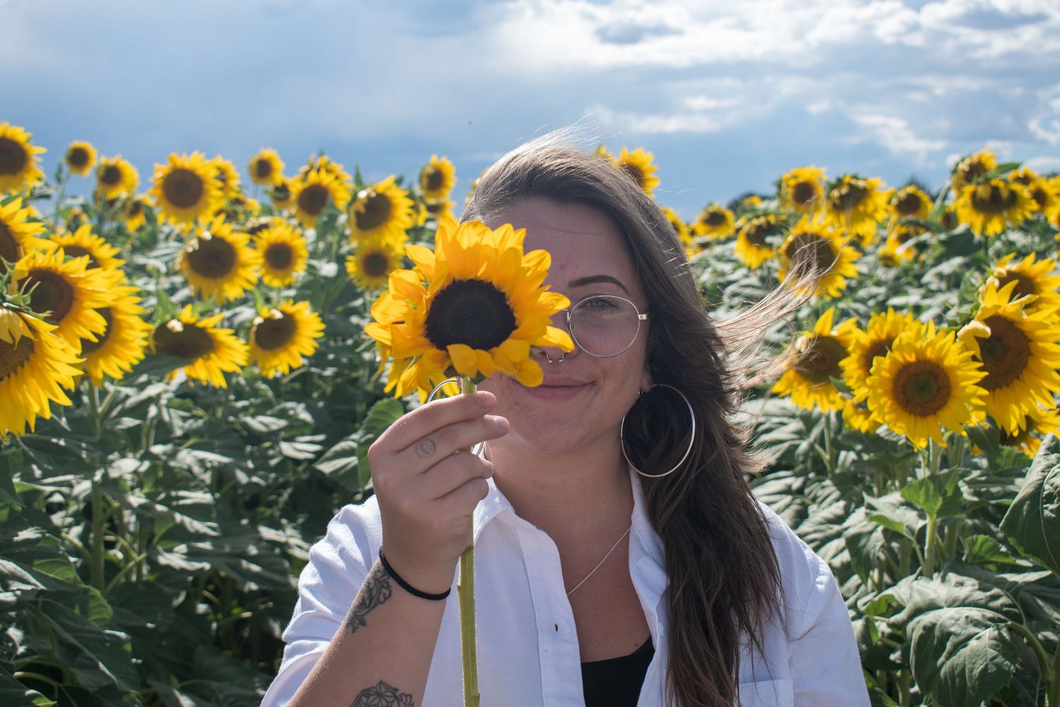 Woman holding a sunflower over her eye while smiling in a vibrant sunflower field under a cloudy sky.