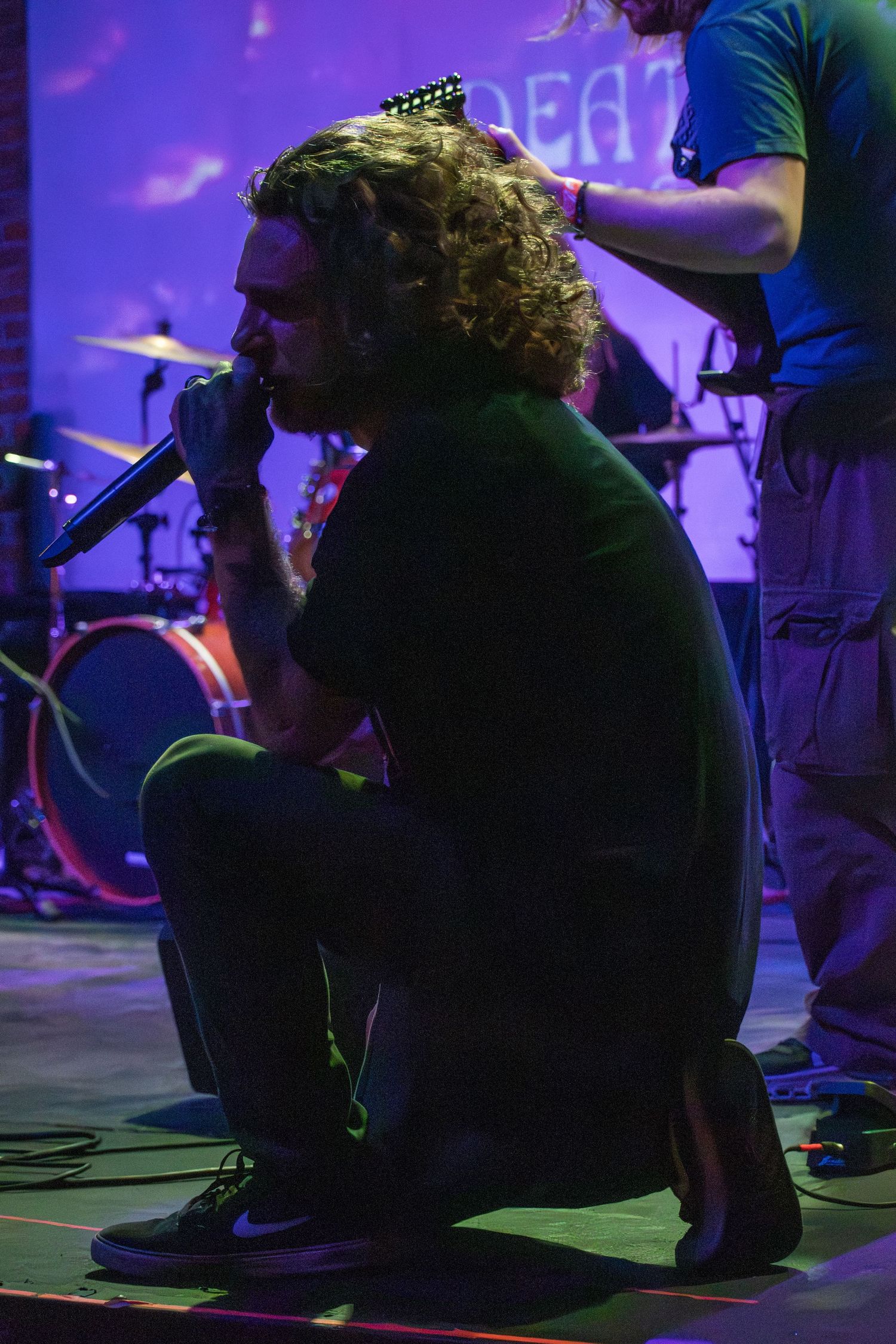 Curly-haired male vocalist crouching on stage singing into microphone at live concert with purple stage lighting.