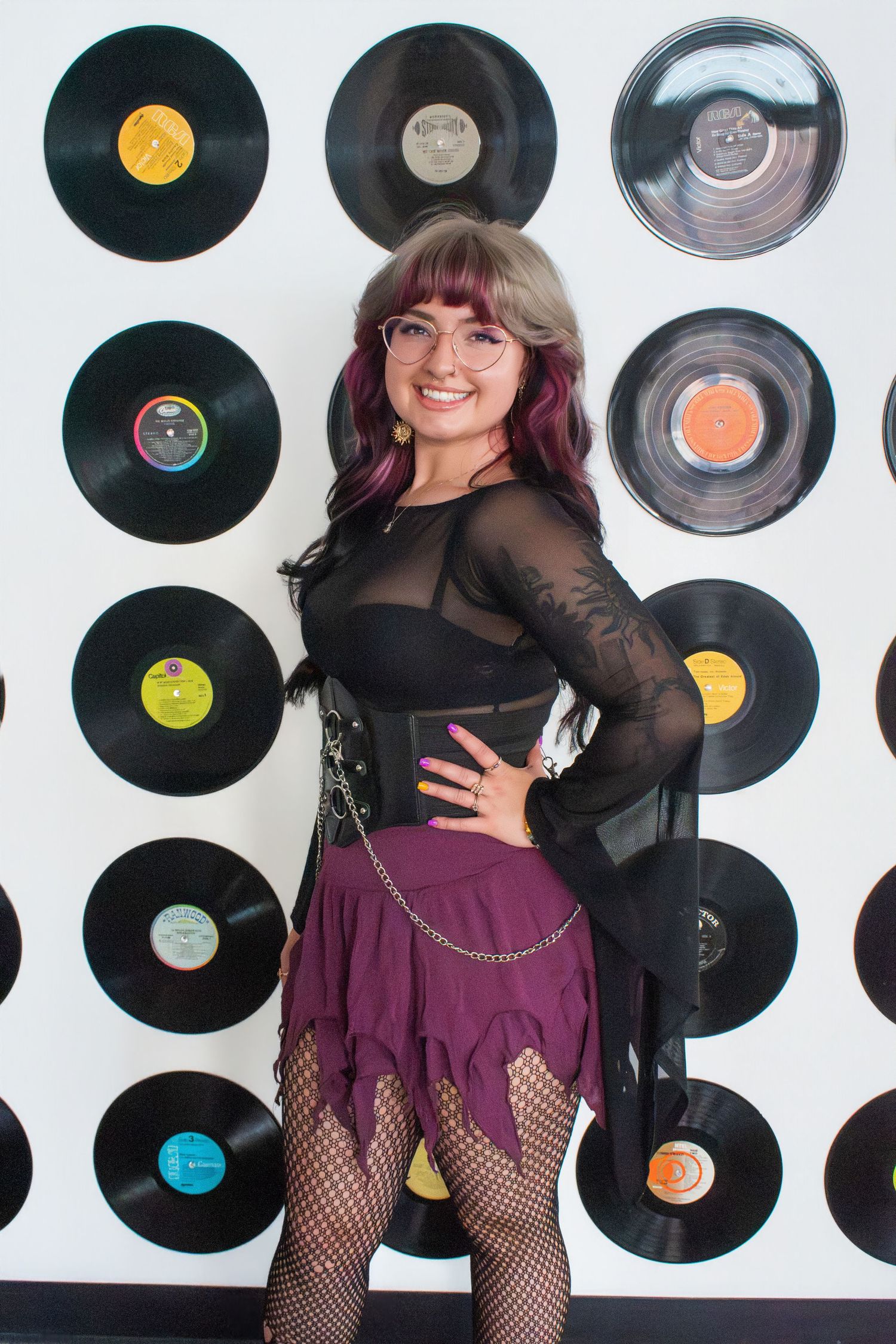 Young woman in purple skirt and fishnet tights poses smiling before a wall of vinyl records.