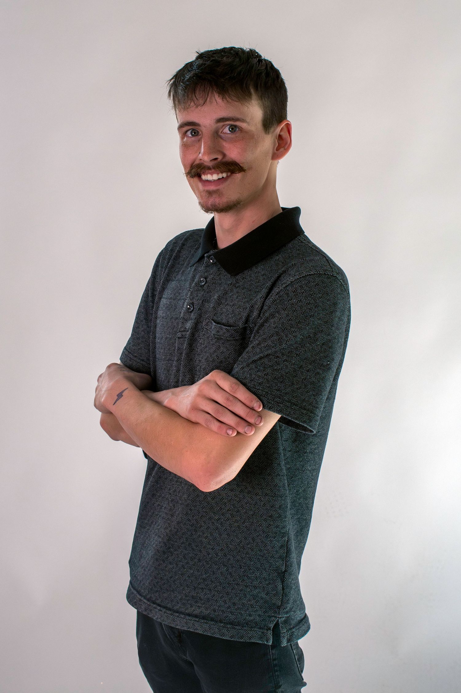 Young man with mustache smiling with arms crossed wearing gray polo shirt against white background.