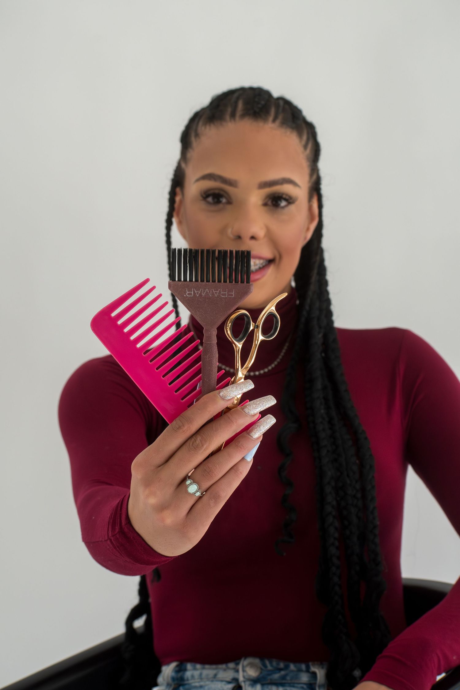 Woman with box braids in burgundy top holds pink comb, brush, and gold scissors toward camera.