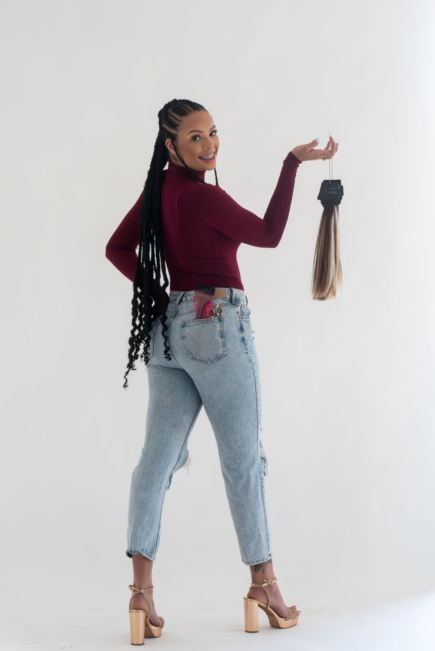 Woman in burgundy top and jeans holding hair extensions, showcasing long braided hairstyle against white background.