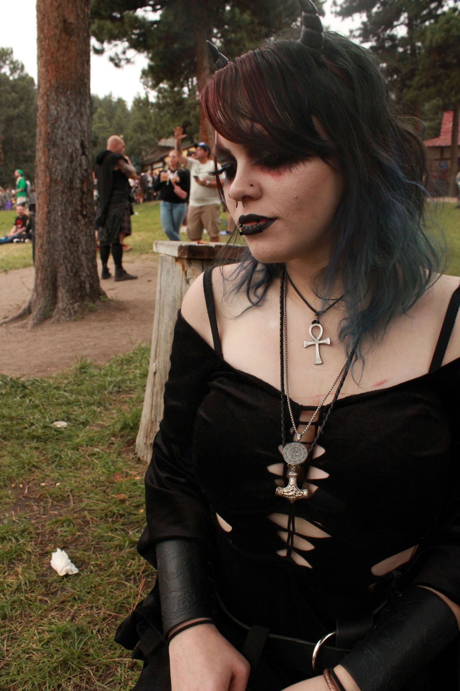 Young goth woman with blue hair and ankh necklace wearing black dress at outdoor festival.