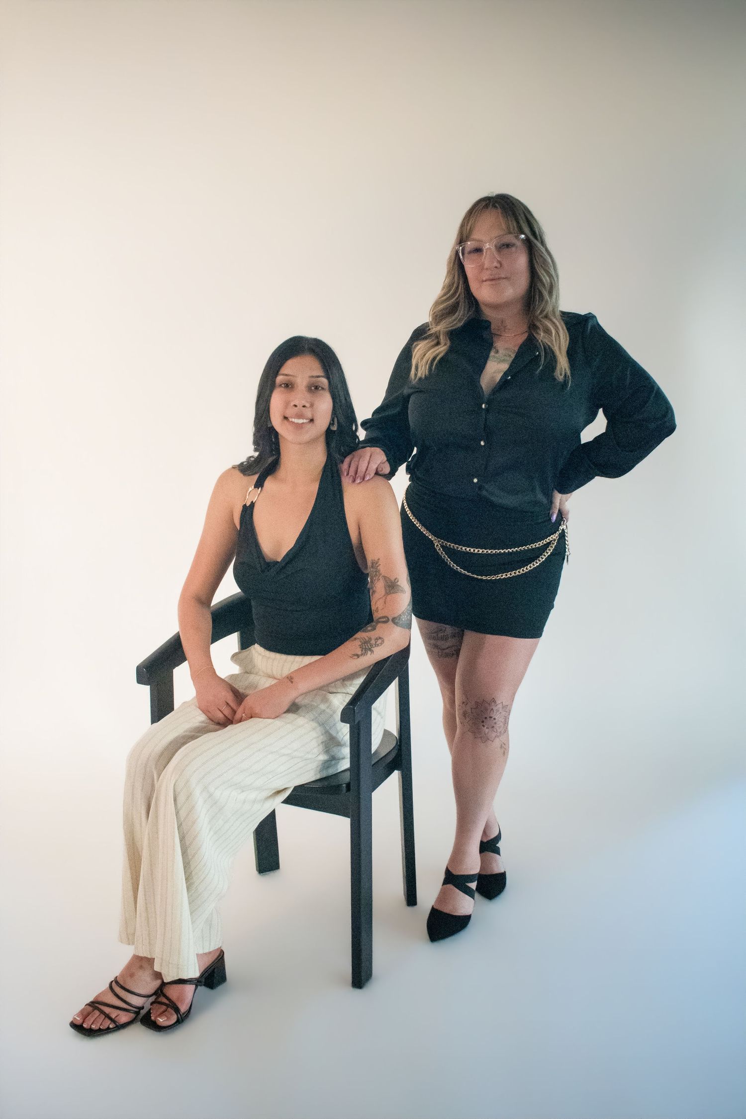Two women posing in a studio, one seated in a black chair wearing white pants, the other standing in a black dress.