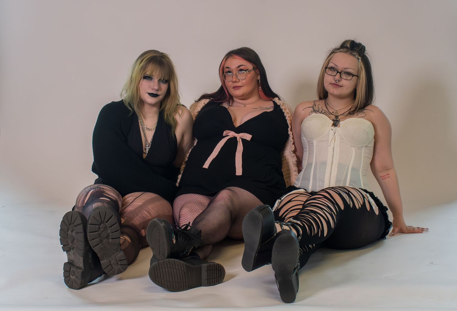 Three young women in gothic fashion sitting together on floor against white background in a studio photo.