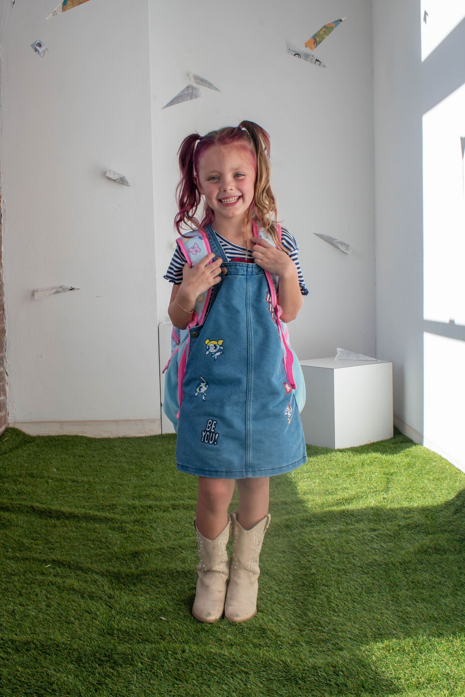 Smiling girl in denim pinafore, striped shirt, cowboy boots, pink backpack, and pigtails stands on green turf.
