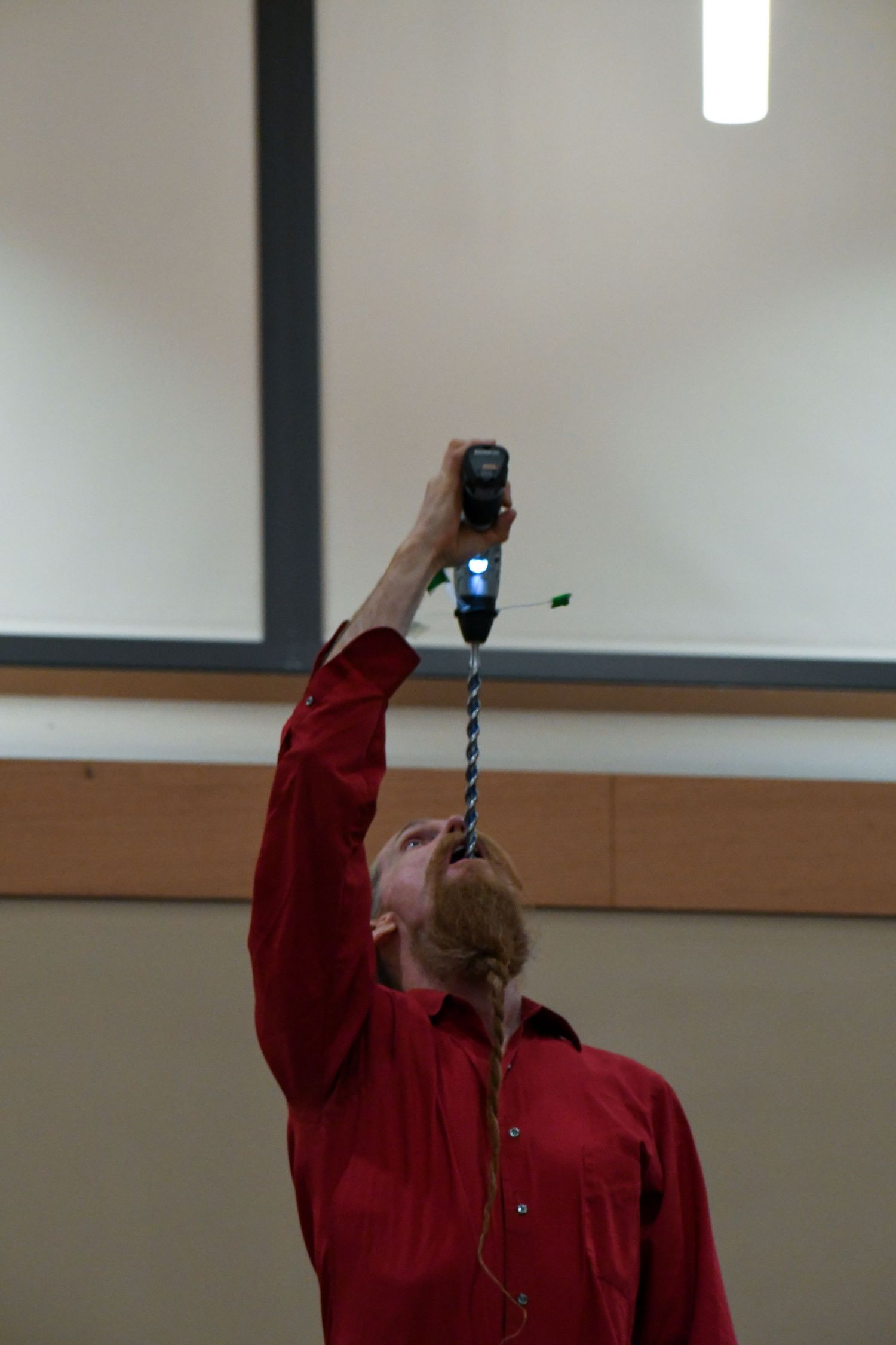 Man in red shirt performing daring stunt with electric drill pointed at his mouth in indoor setting.