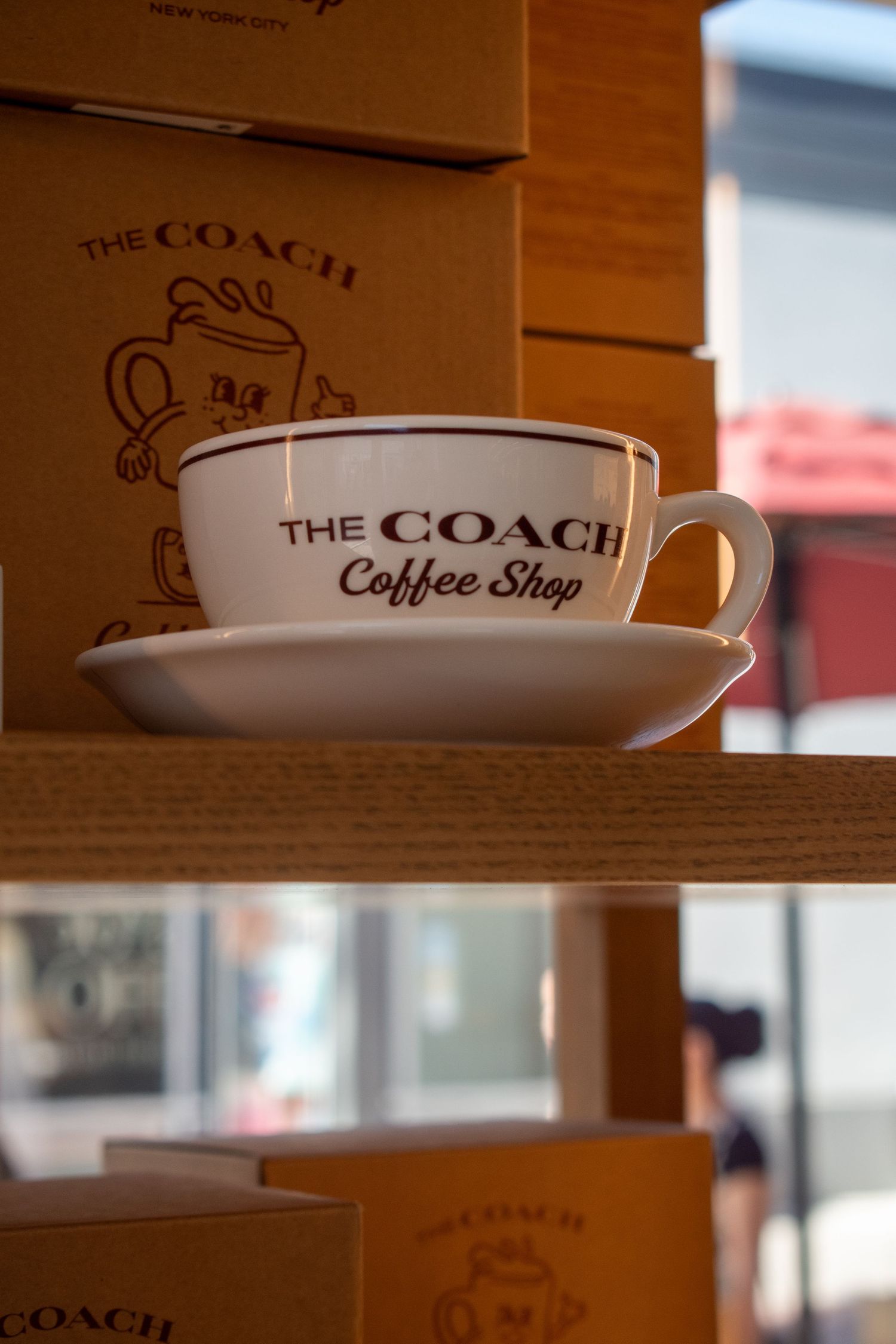 White ceramic cup and saucer branded The Coach Coffee Shop displayed on a wooden shelf with matching gift boxes.