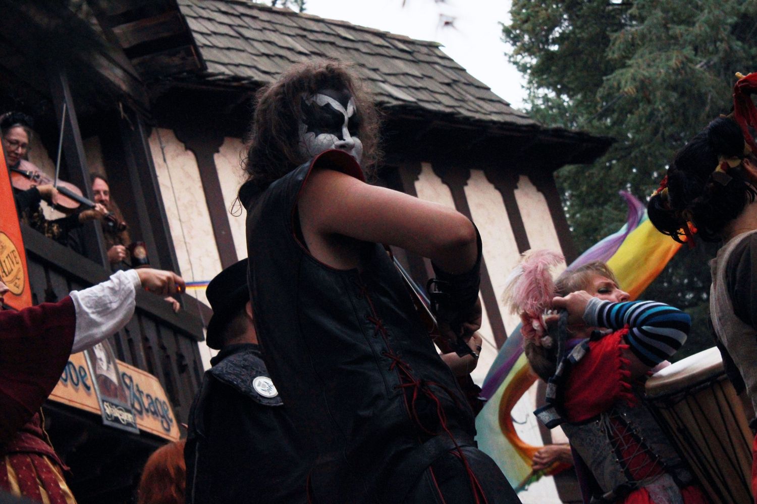 Person in dark costume with face paint performs at a Renaissance fair with colorful flags in background.