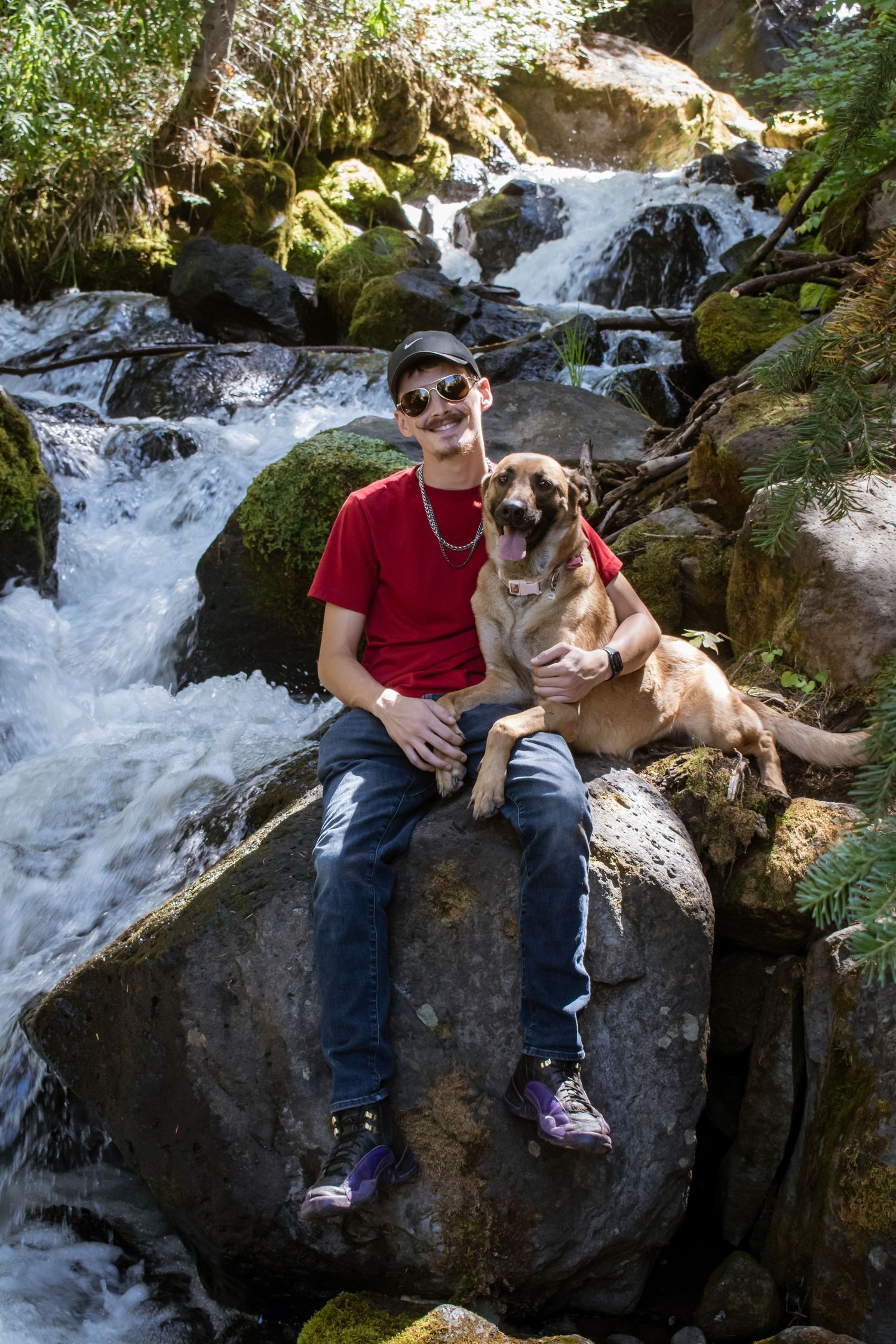 Young man sitting on rocks with his happy dog in front of a scenic waterfall.
