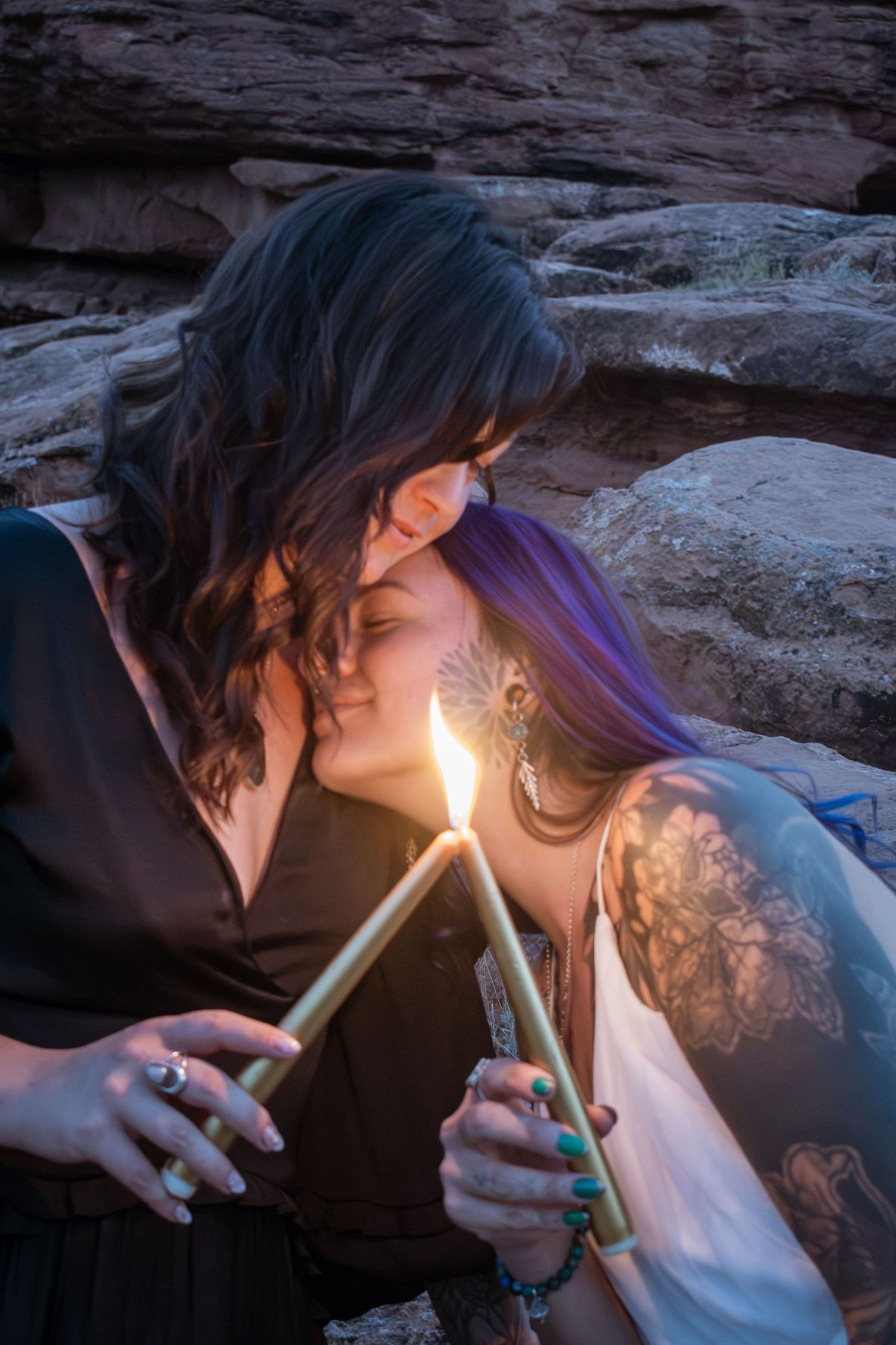 Two women share an intimate moment holding a lit candle together among rocky formations at dusk.