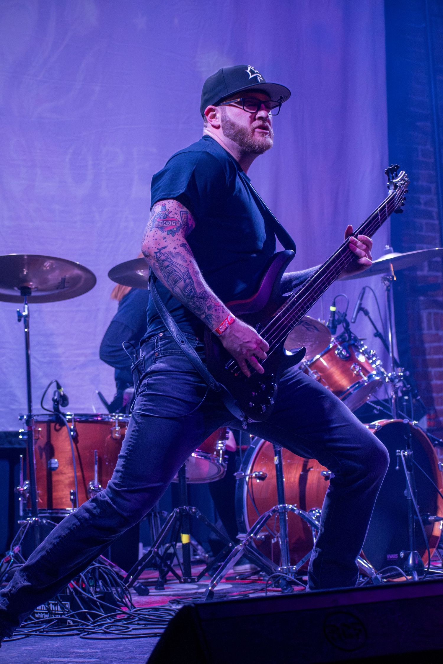 Tattooed male guitarist performing on stage under purple lights with drummer visible in background at live concert.