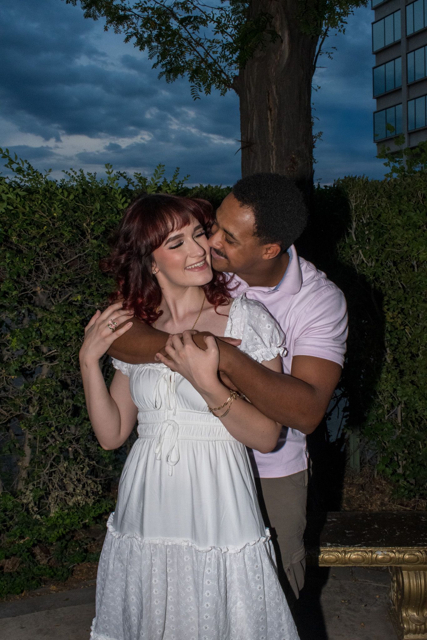A couple shares a romantic embrace outdoors, man in pink shirt kissing woman in white dress with red hair at dusk.