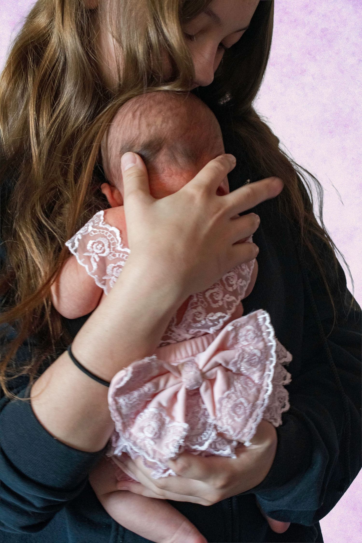 A young woman tenderly holds and kisses a newborn baby dressed in a delicate pink lace outfit.