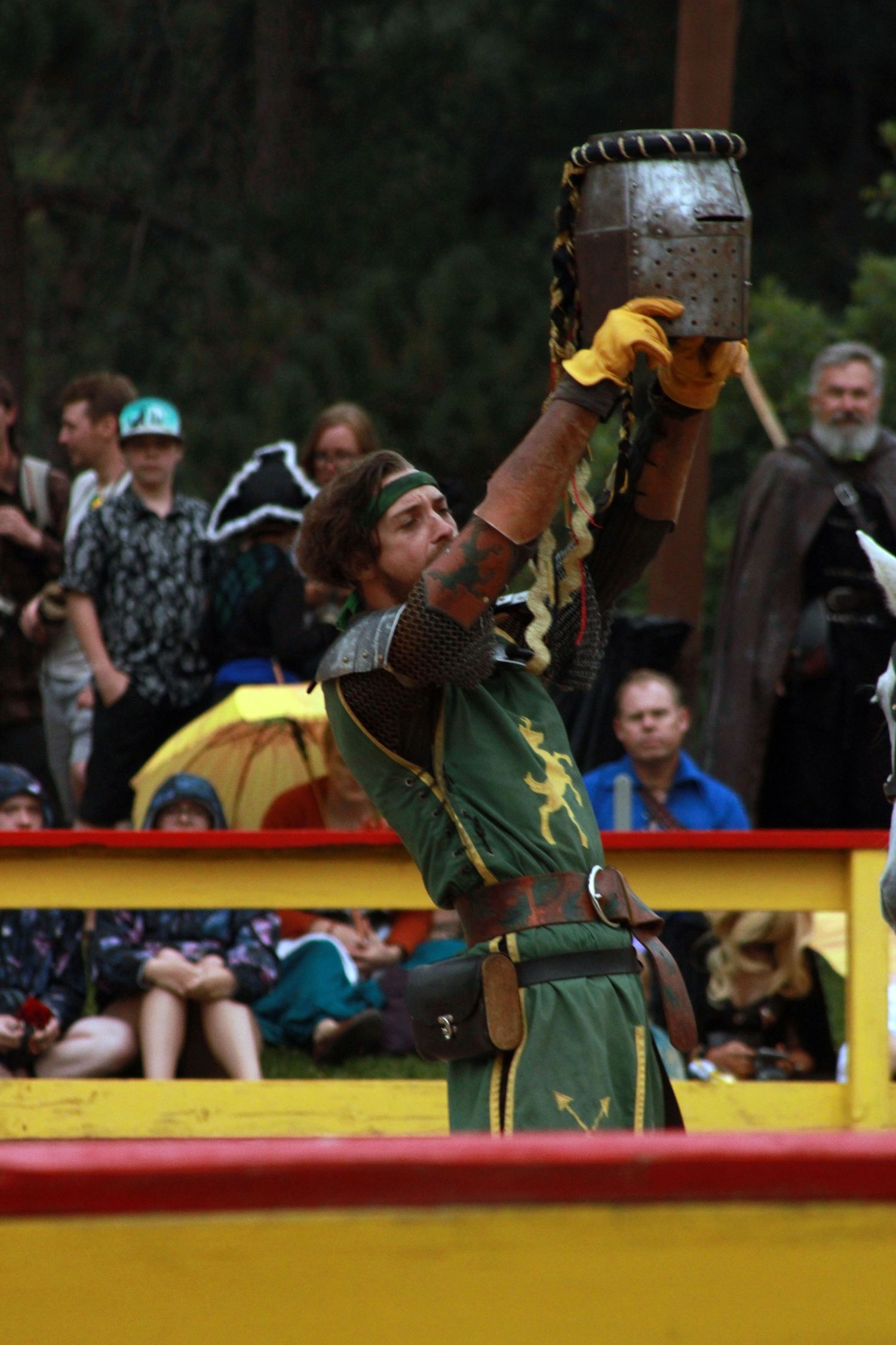 Medieval performer in green costume raises a helmet at a Renaissance faire while spectators watch from the stands.