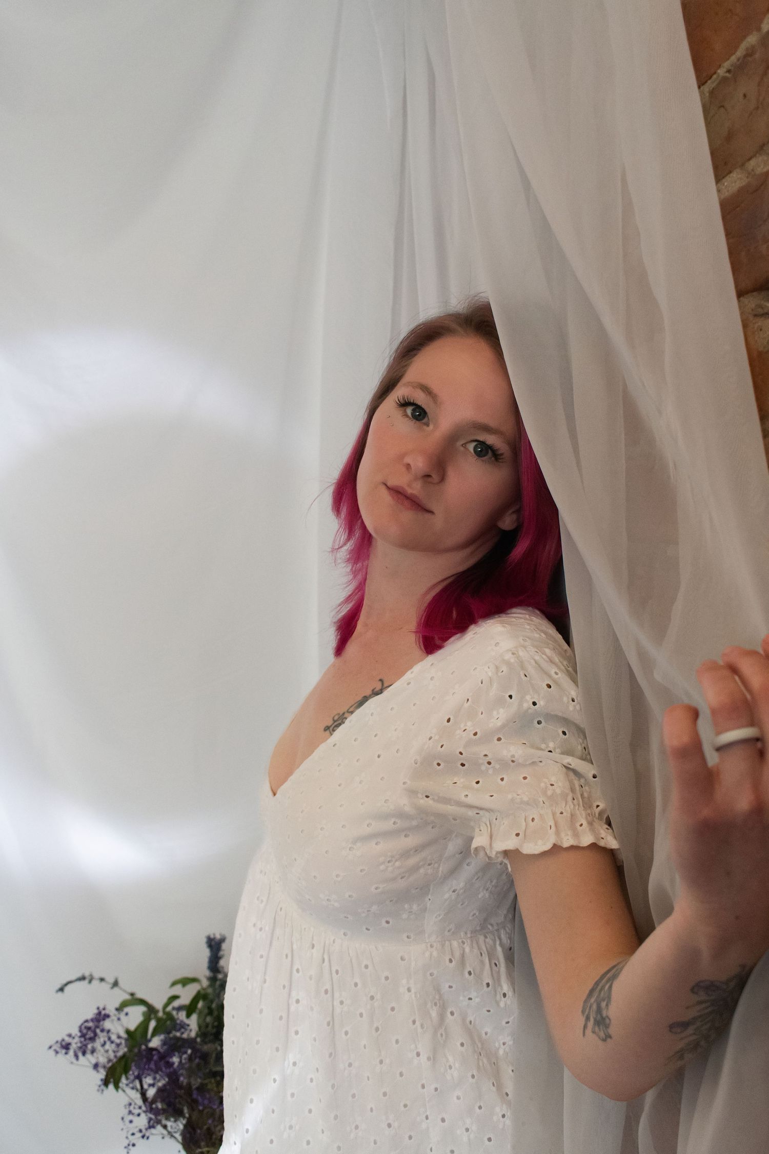 A young woman with pink hair in a white eyelet dress peers through sheer white curtains, with a tattoo visible on her arm.
