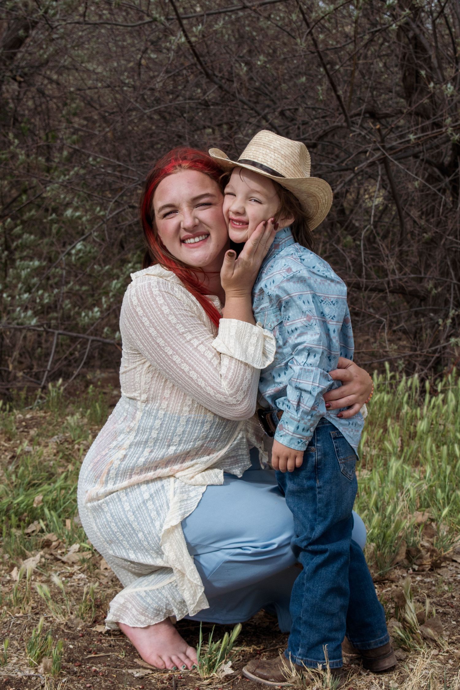 A smiling red-haired woman crouches outdoors hugging a young boy in a cowboy hat and blue western shirt.