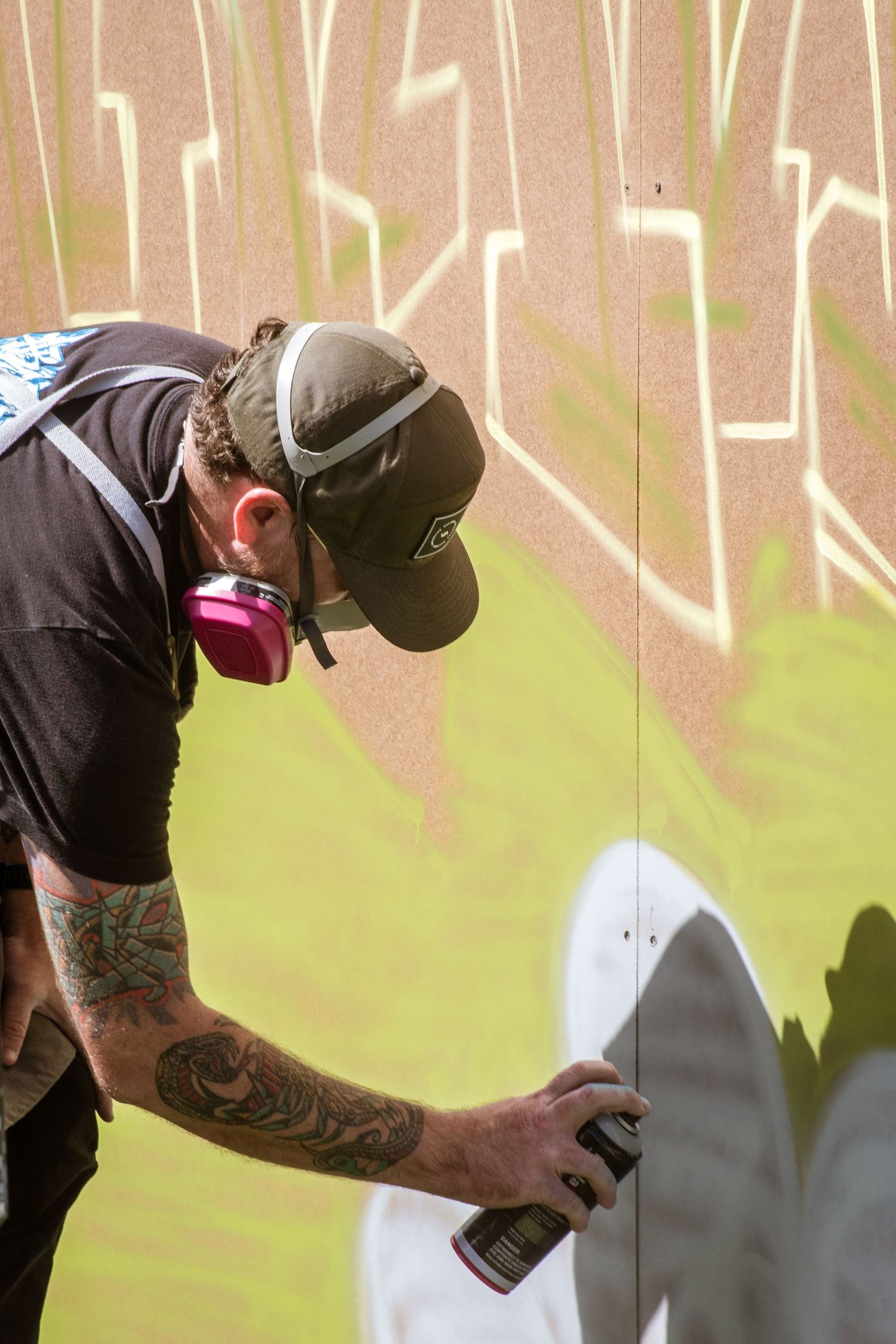 Tattooed graffiti artist wearing cap and respirator sprays colorful street art on a wall outdoors.