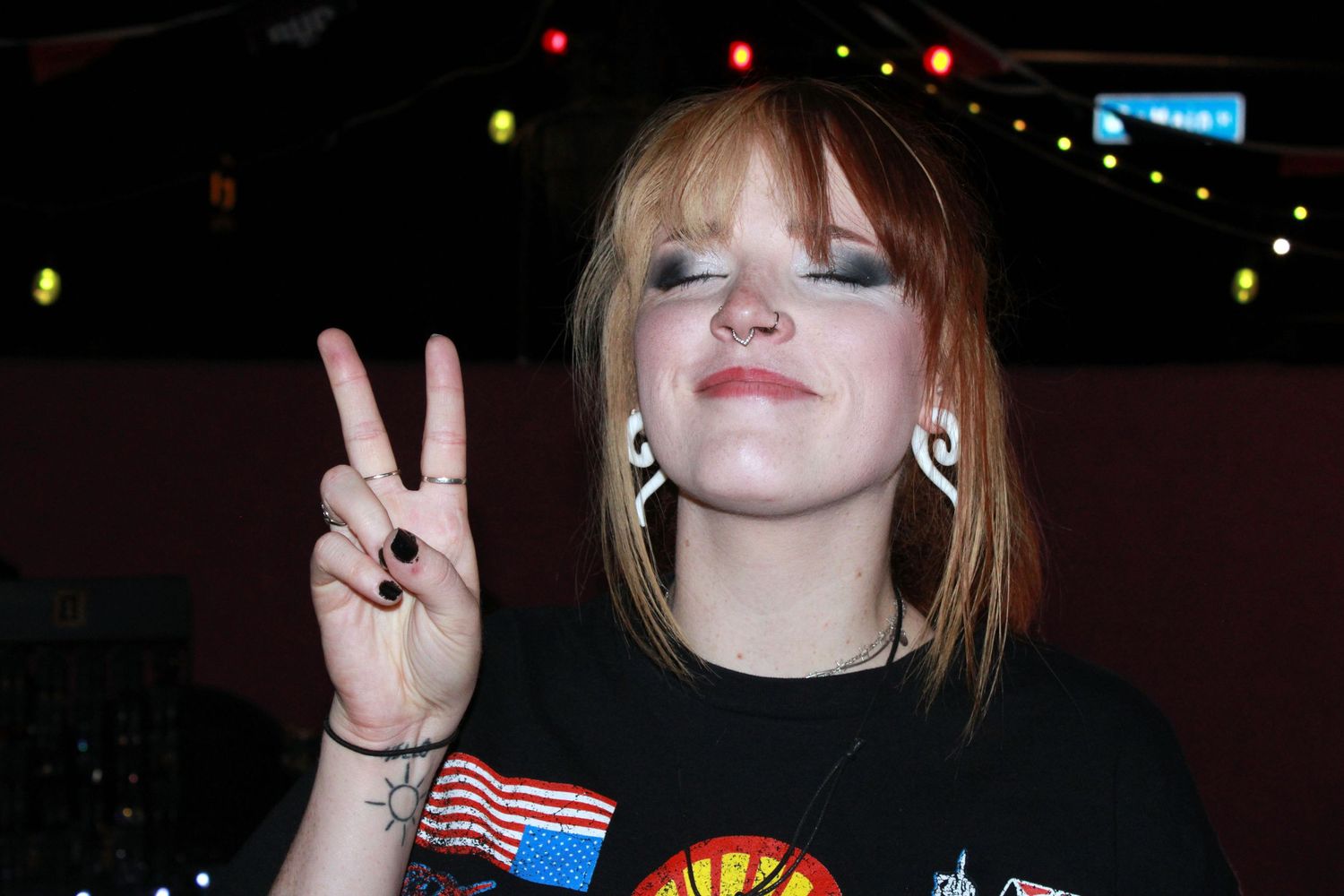 Young woman with red hair making a peace sign gesture, wearing a black graphic tee in a dimly lit venue.