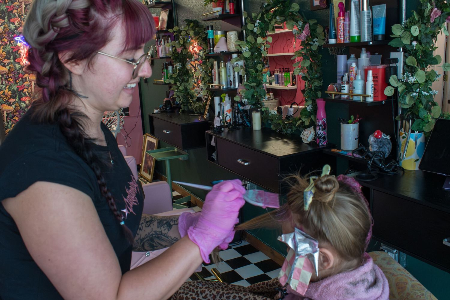 Hair stylist in pink gloves working on a young girl's hair in a colorful salon with product shelves.