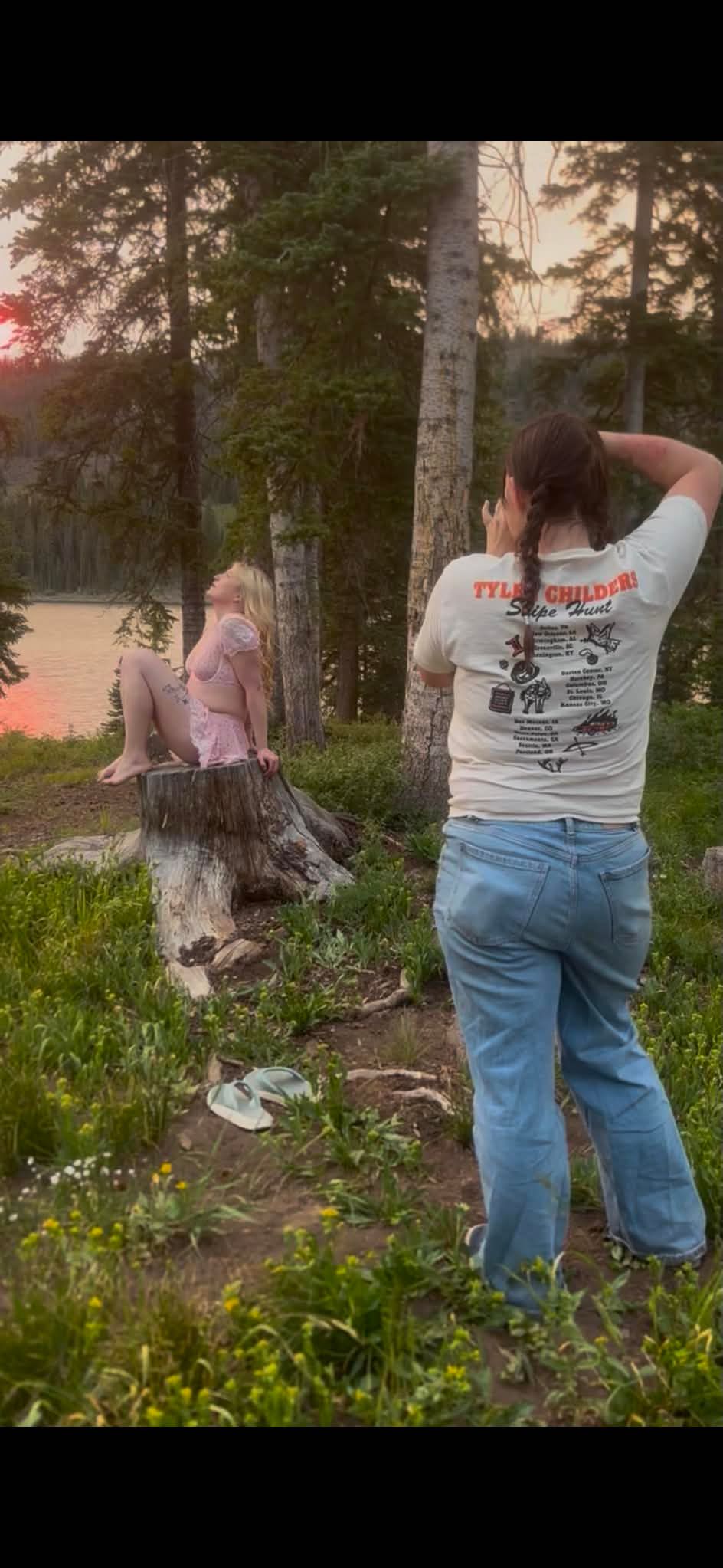 Photographer in Tyler Childers shirt capturing woman posed on tree stump at lakeside sunset.