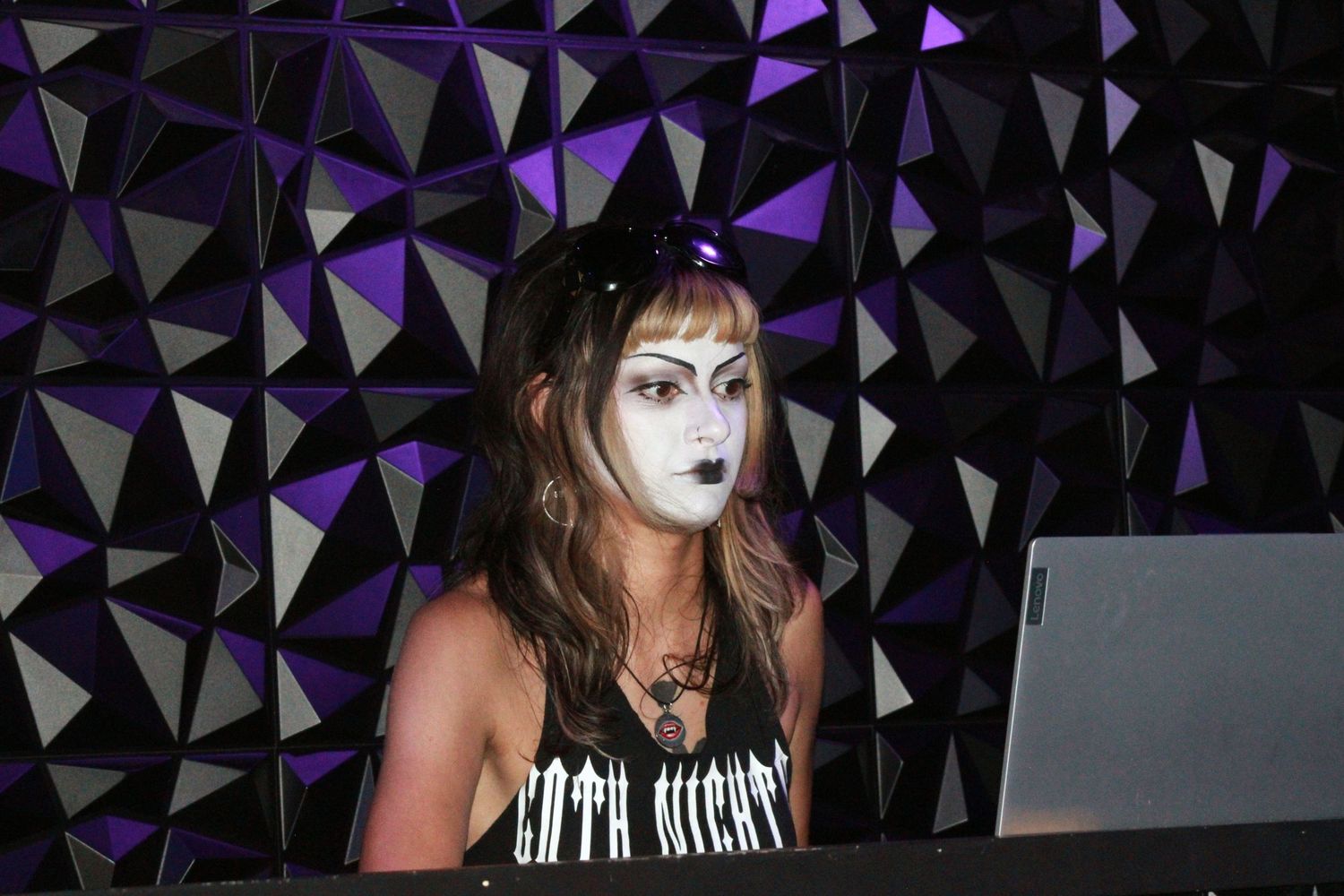 Person in gothic makeup with white face paint performing as a DJ at a purple-lit event.