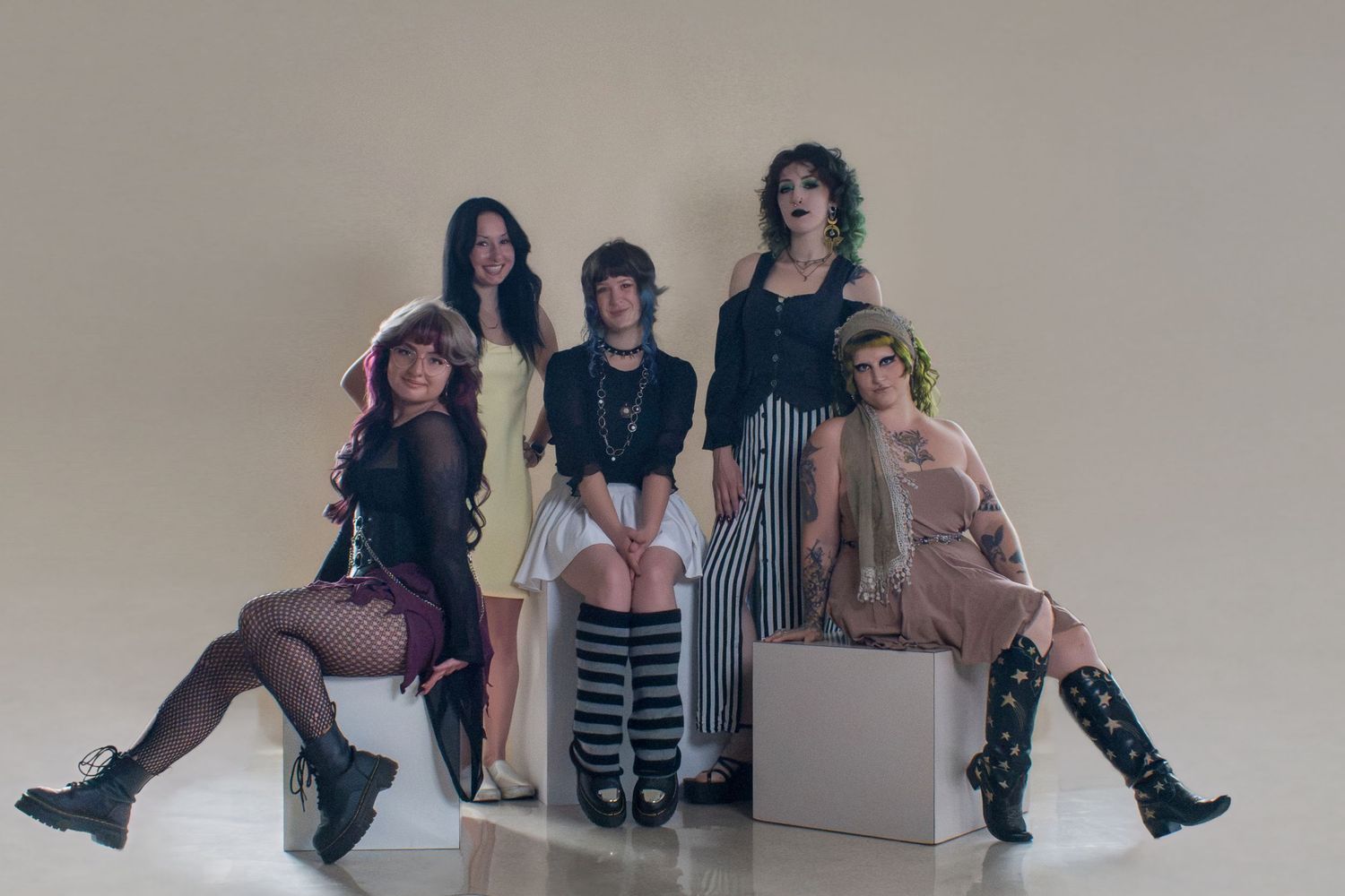 Five stylish young women pose together in a studio, wearing edgy fashion outfits with boots and dark accessories.