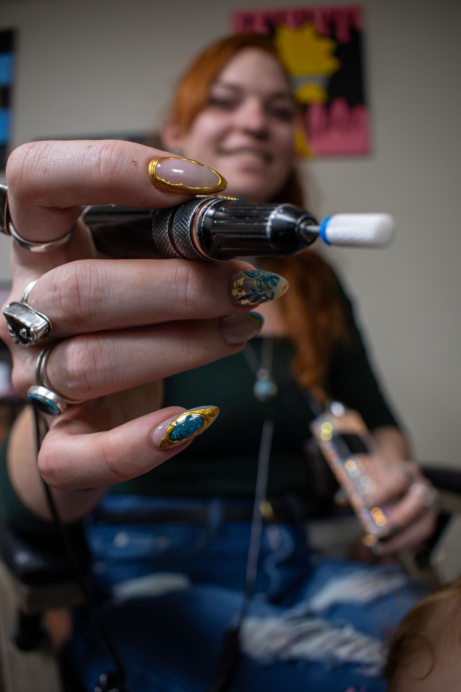 Tattoo artist with decorative nails holds a tattoo machine toward camera, smiling in a tattoo studio.