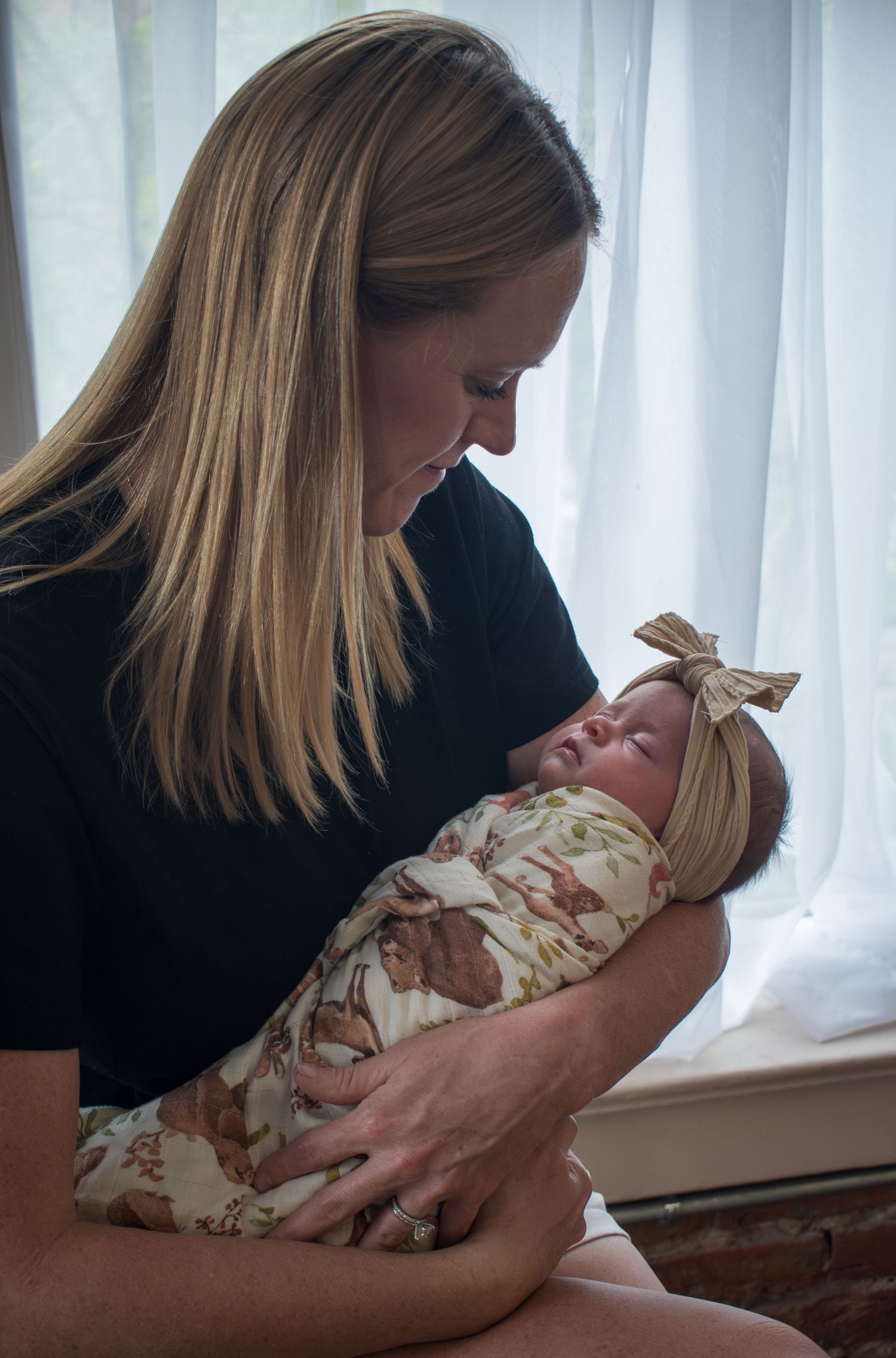 A mother tenderly gazes at her newborn baby wrapped in a floral swaddle with a bow headband by a bright window.