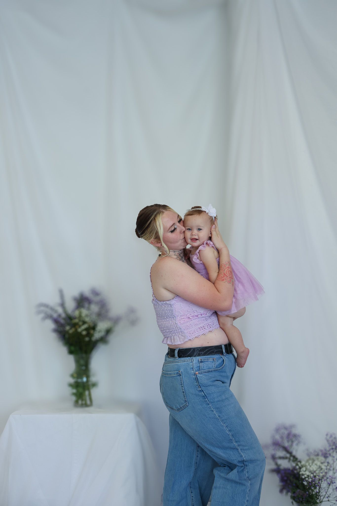 Mother in pink crop top and jeans kisses baby girl in pink tutu against white draped backdrop.