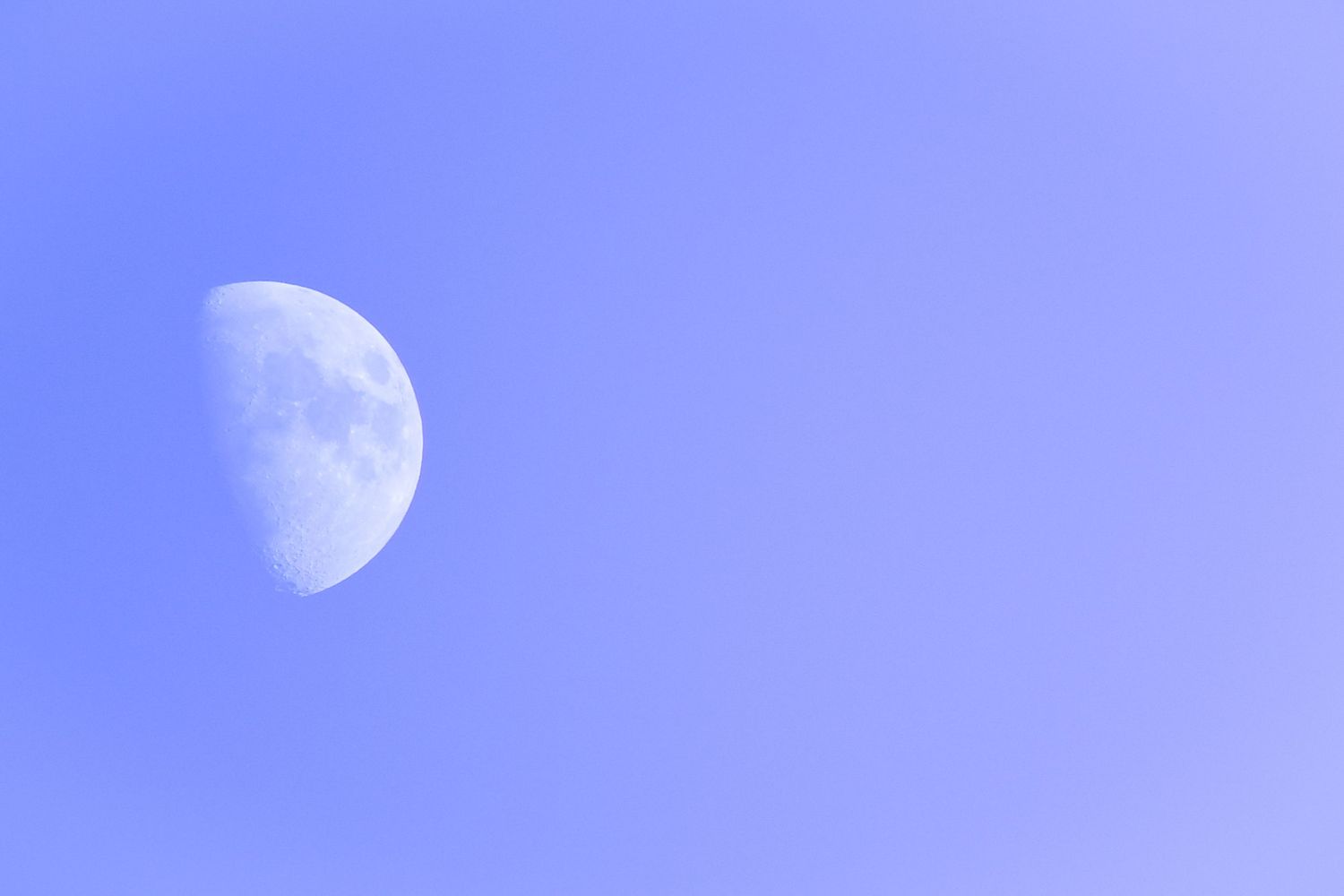 Half moon visible in clear blue daytime sky with soft purple hue.
