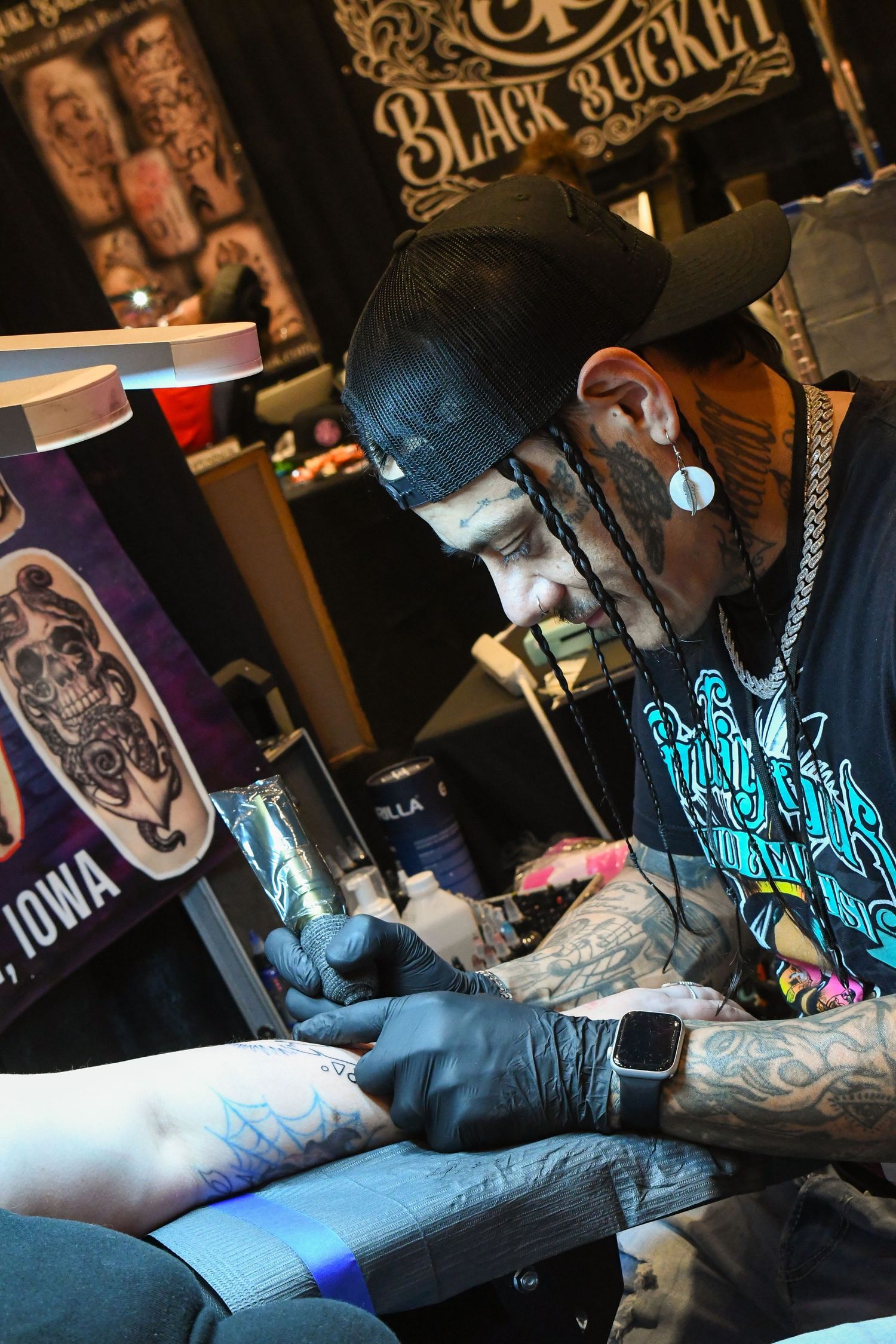 Heavily tattooed tattoo artist working at Black Bucket booth, wearing black cap and gloves while tattooing a client.