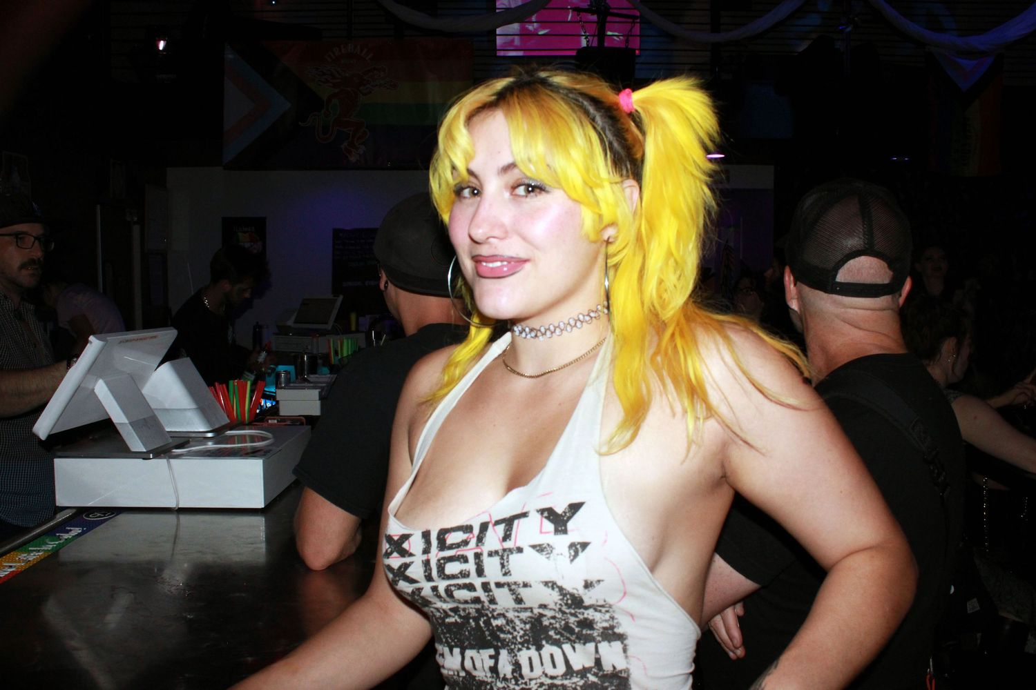Woman with bright yellow pigtails wearing a graphic tank top poses at a bar in a nightclub setting.