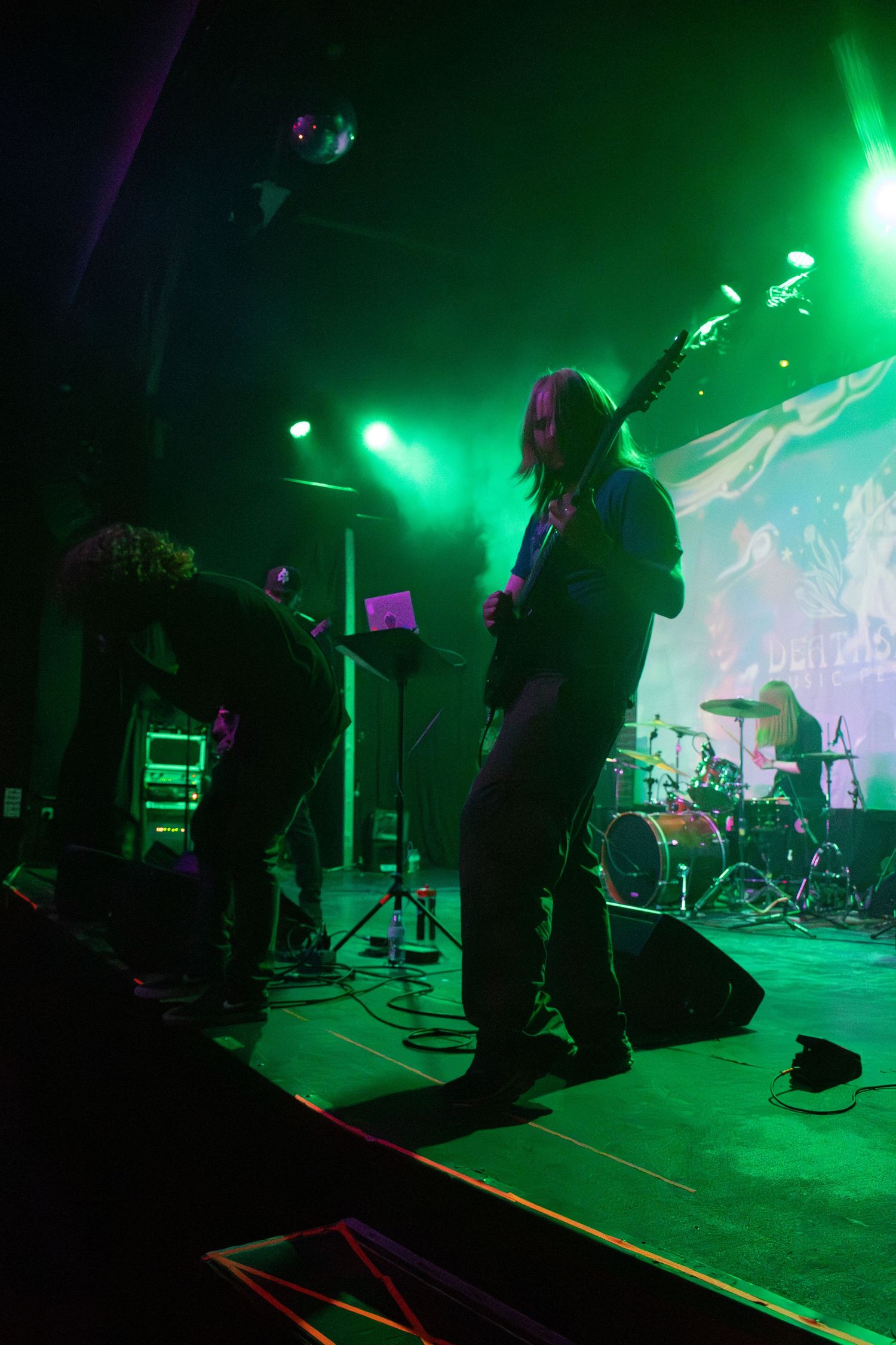 Band performing on stage under green lights, guitarist playing electric guitar with drummer visible in background.