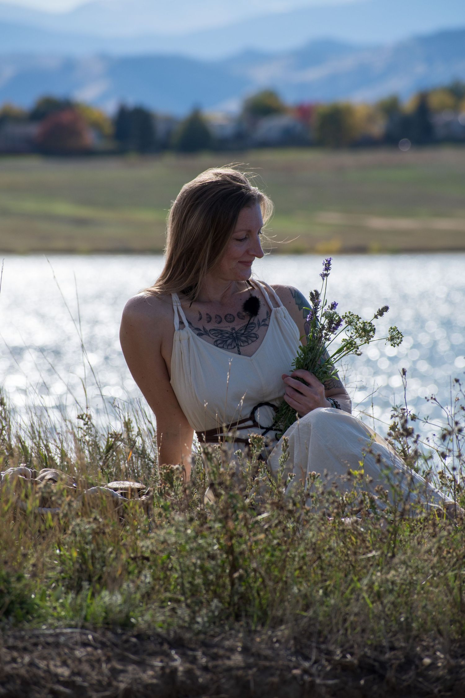 Woman sitting by a lake holding wildflowers, wearing a white dress with tattoos, mountains in the background.