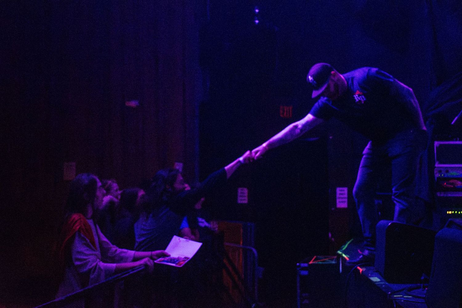 Performer leans toward audience on dimly lit stage bathed in purple and blue light during live concert.