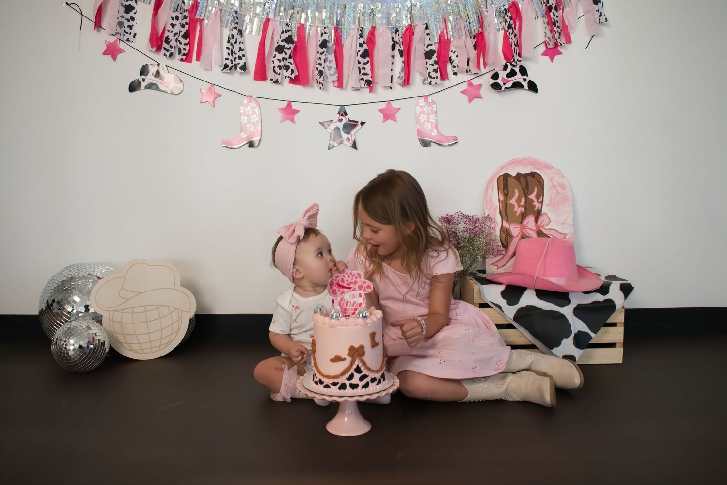 Two girls celebrate a first birthday with a pink cake and cowgirl-themed decorations including banners and stars.