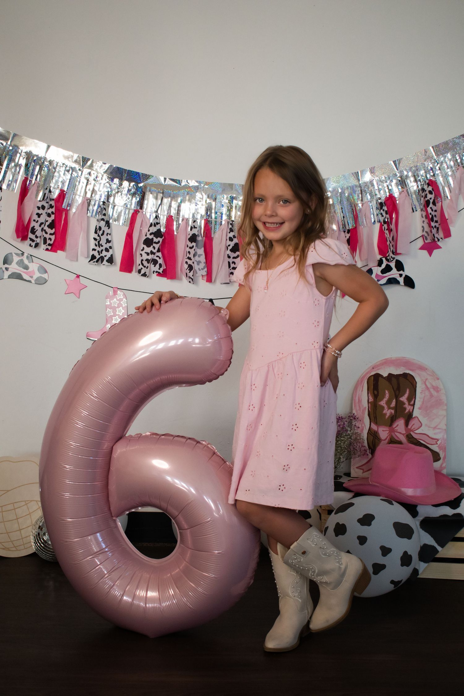 Smiling girl in pink dress holding large number 6 balloon at cowgirl themed birthday party.