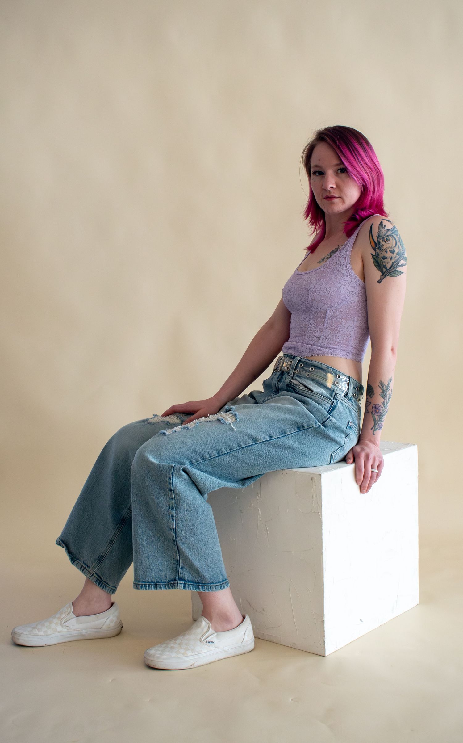 Woman with pink hair and tattoos sits on white cube in jeans and purple crop top.