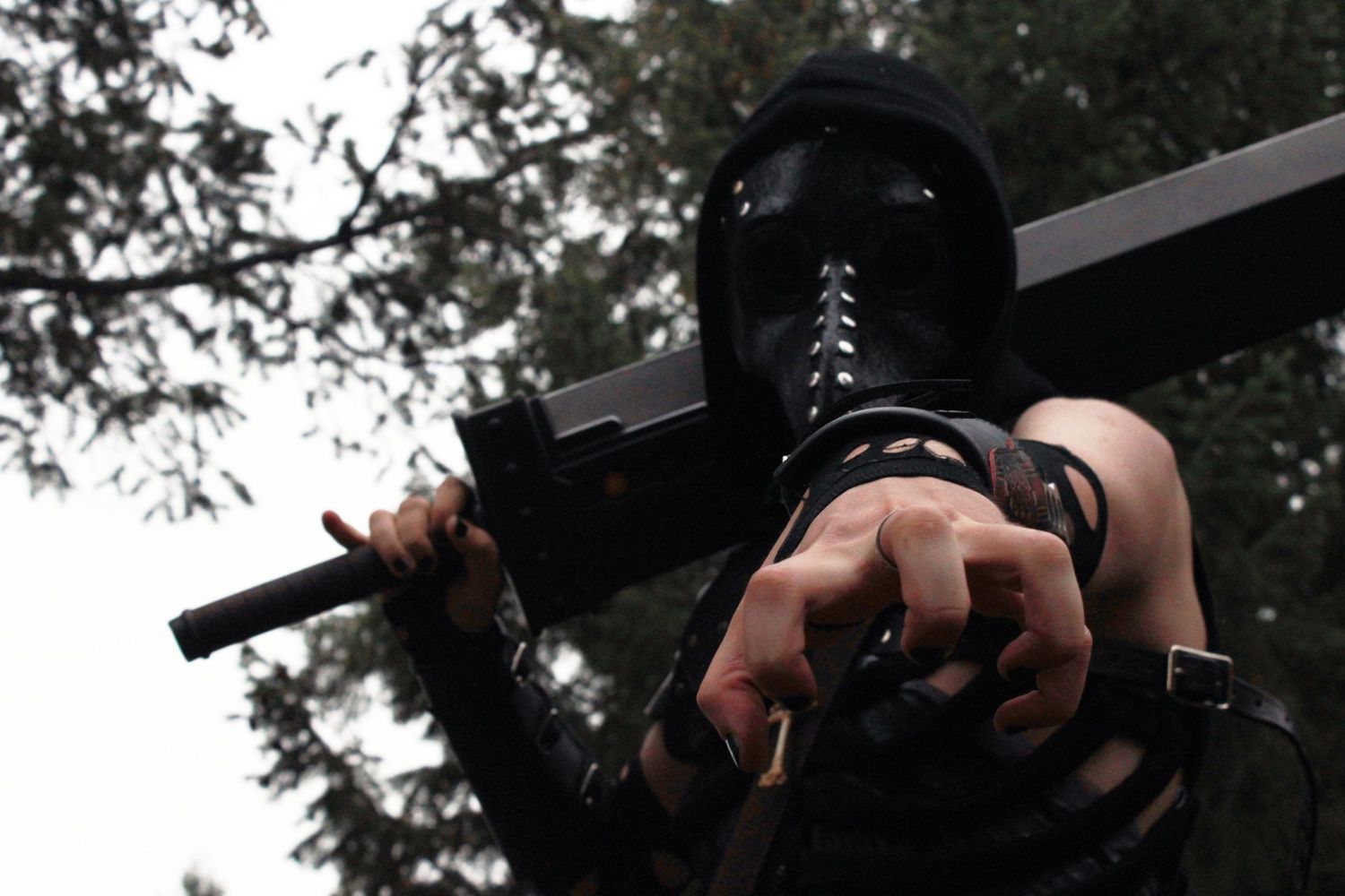 Person in black mask and tactical gear holding a weapon outdoors among trees.