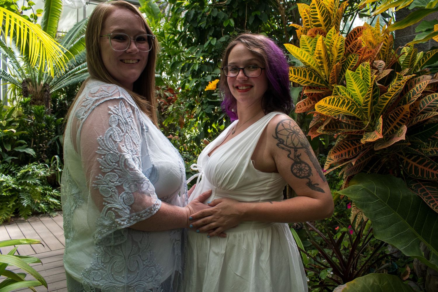 Two women in elegant dresses pose together in a lush tropical garden setting.