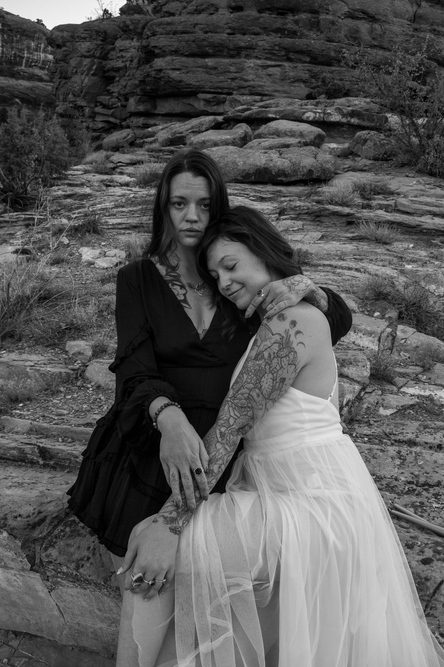 Two tattooed women in black and white photo, one in white dress, embracing on rocky desert landscape.