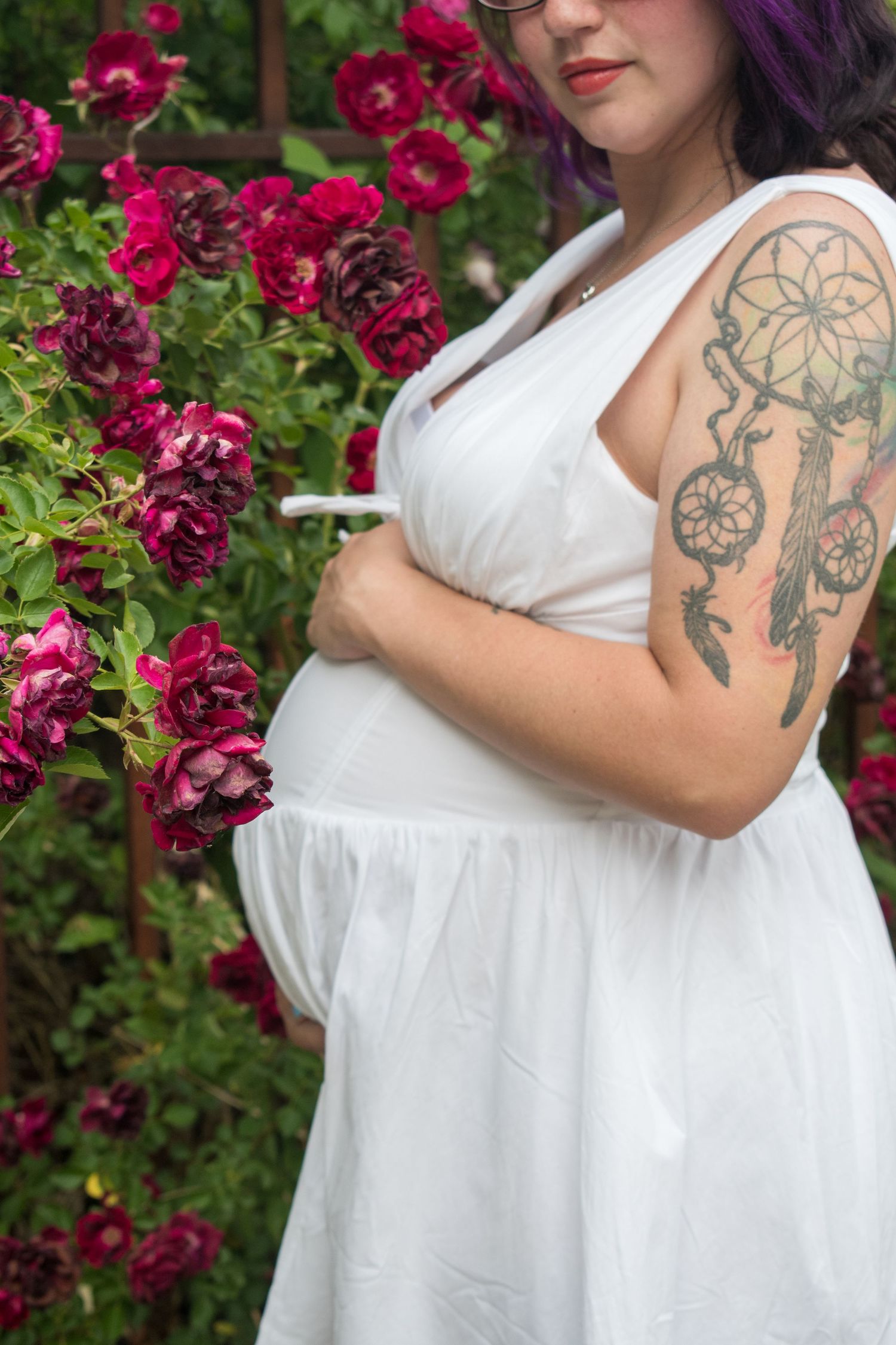Pregnant woman in white dress with dreamcatcher tattoo standing beside red roses in a garden.
