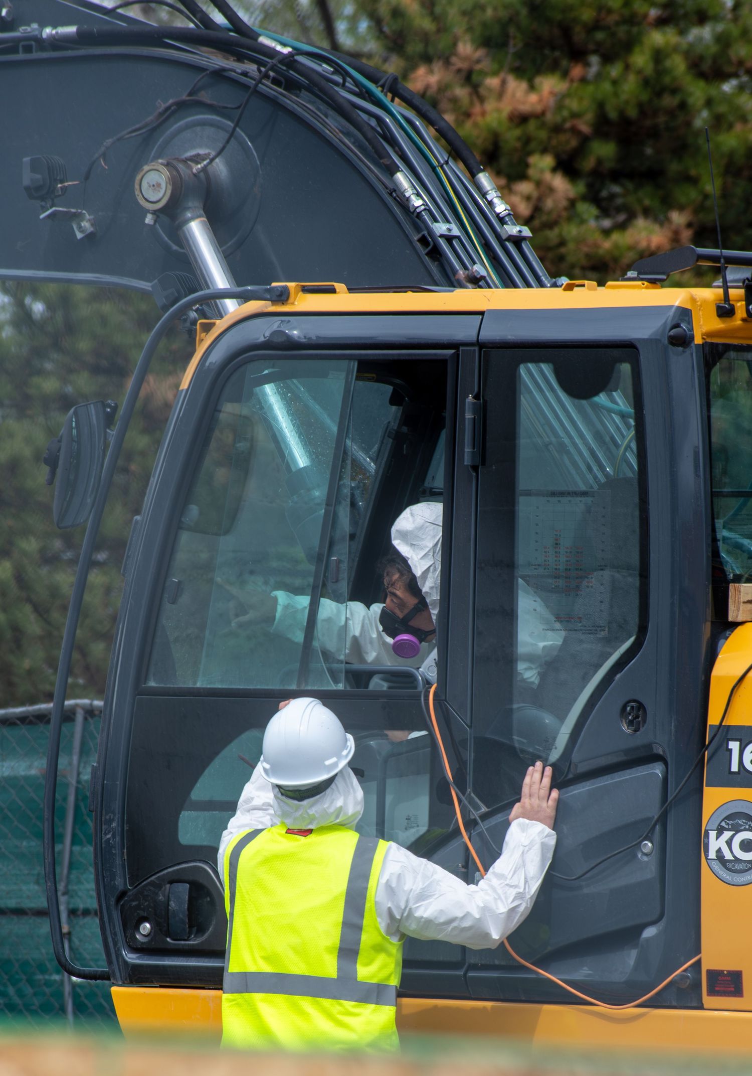 Construction worker in hazmat suit operating yellow excavator while colleague in hi-vis vest signals directions on site.