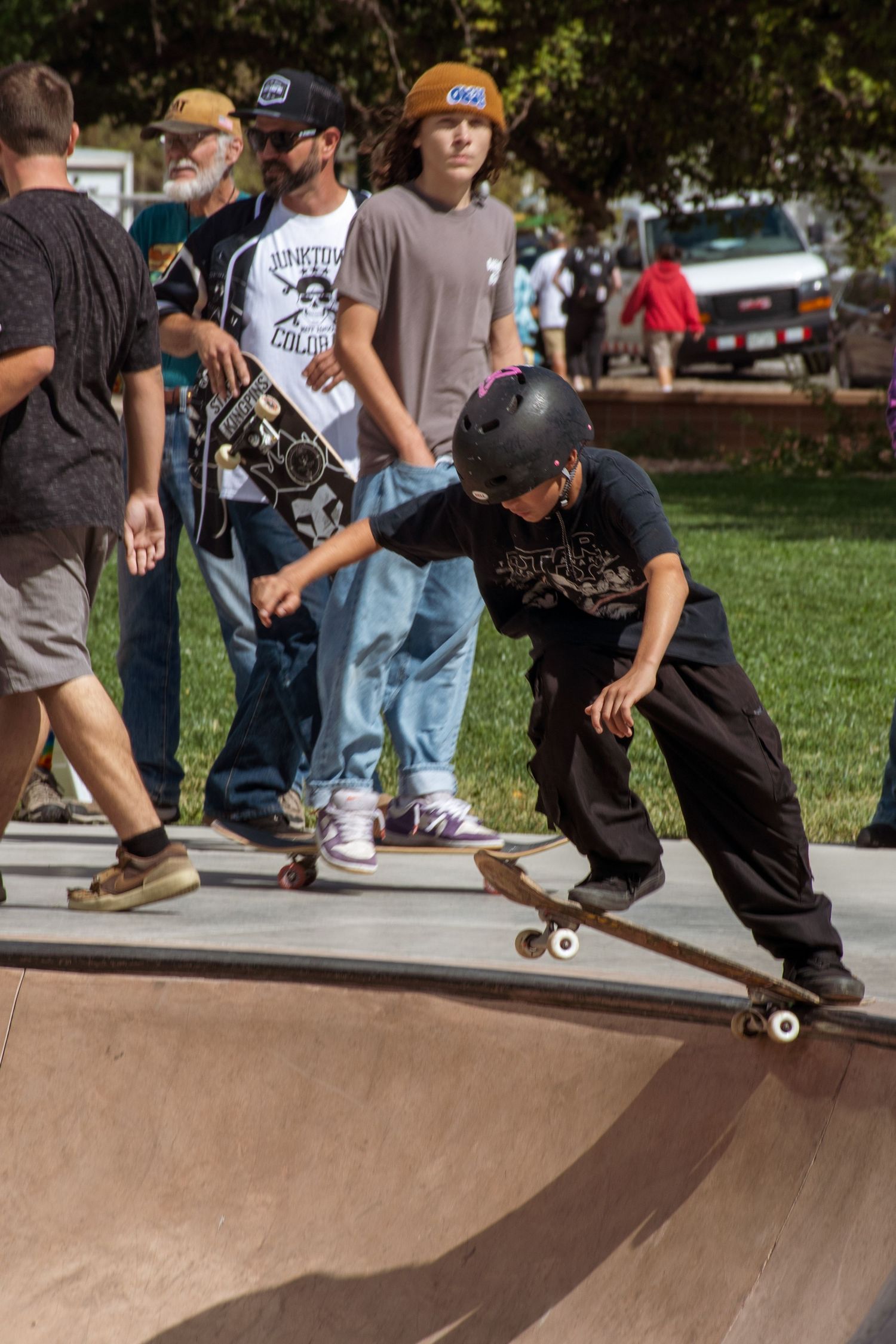 Young skateboarder in helmet performs trick on ramp at outdoor skate park while spectators watch in background.