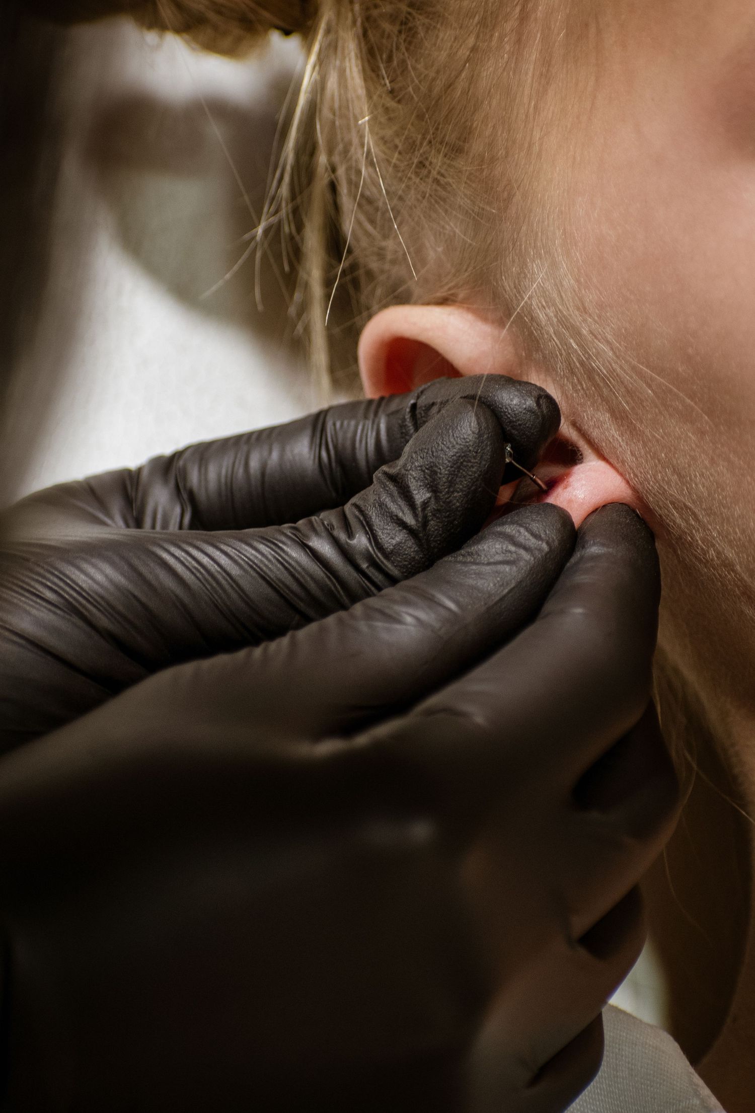 Professional piercer wearing black gloves carefully performing an ear piercing procedure on a blonde client.