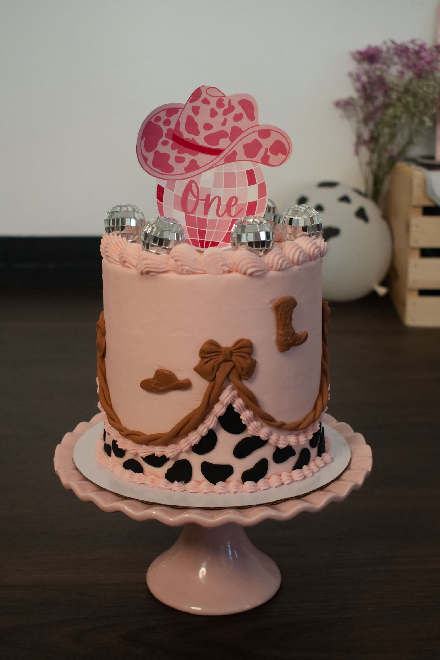 Pink cowgirl first birthday cake with disco balls, cow print, bow details, and a pink hat topper reading One on a cake stand.